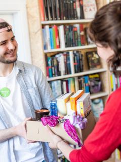 Der ehrenamtliche Mitarbeiter Jeremias M. nimmt im Oxfam Shop in Halle/Saale einen Karton mit Sachspenden von einer Sachspenderin (hier: Hanna Charlotte Boehme) entgegen. Eine Frau übergibt einem Mann einen Karton mit Büchern, Halstüchern und anderen Dingen.