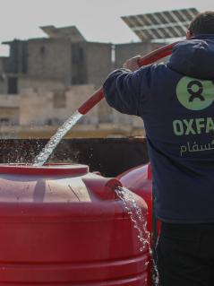 Ein_e Mitarbeiter_in von Oxfam in Syrien befüllt einen Wassertank, damit die Menschen in Notunterkünften Zugang zu sauberem Trinkwasser haben. Ein_e Oxfam-Mitarbeiter_in, mit einer blauen Kaputzenjacke mit grünem Oxfam-Logo bekleidet, befüllt mithilfe eines Schlauches einen roten Wassertank mit sauberem Trinkwasser.