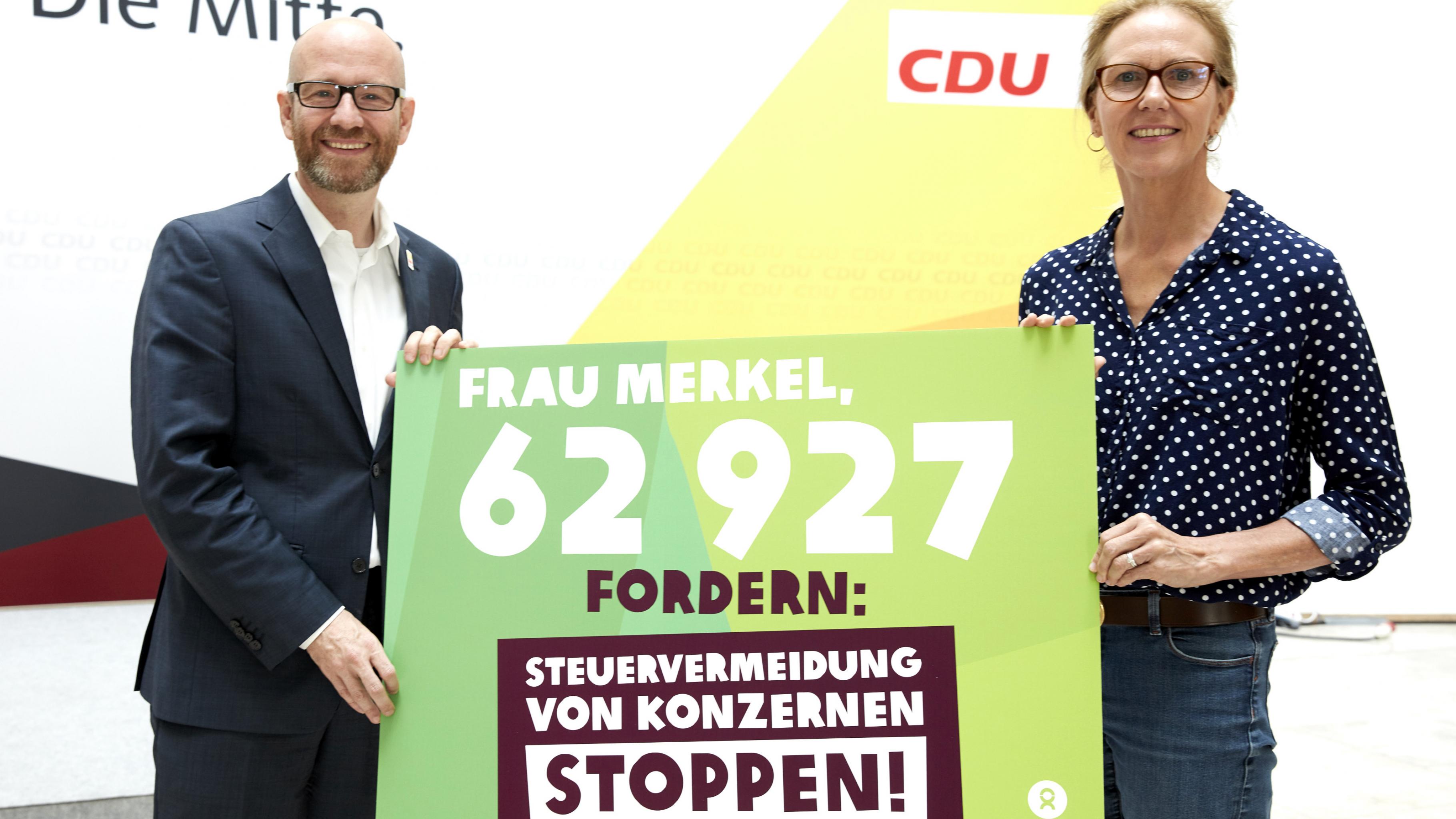Marion Lieser und Peter Tauber halten ein Plakat mit der Aufschrift: „Frau Merkel, 62927 fordern: Steuervermeidung von Konzernen stoppen!“