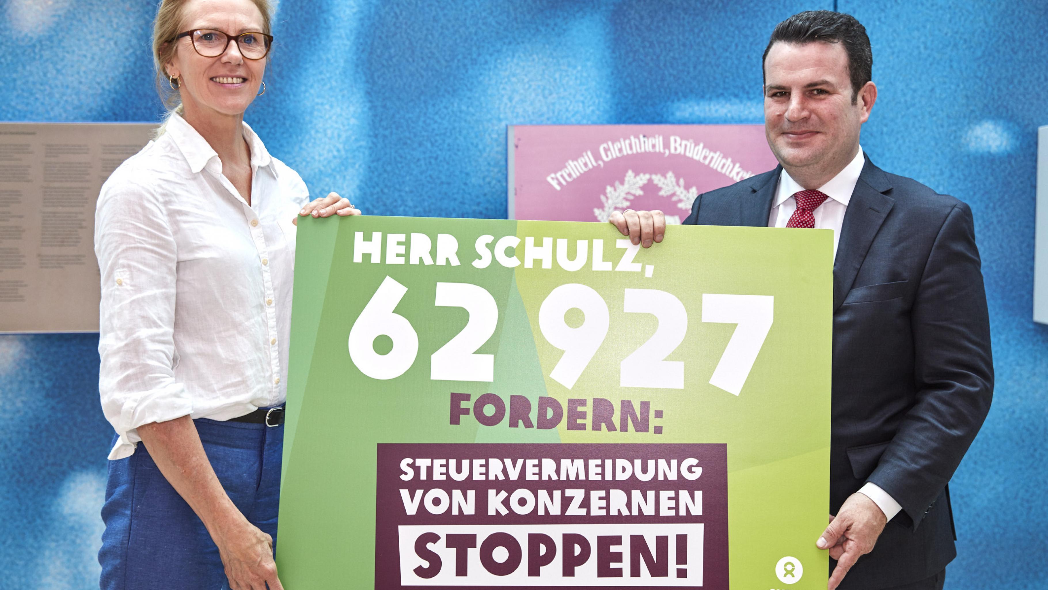 Marion Lieser und Hubertus Heil halten ein Plakat mit der Aufschrift: „Herr Schulz, 62927 fordern: Steuervermeidung von Konzernen stoppen!“