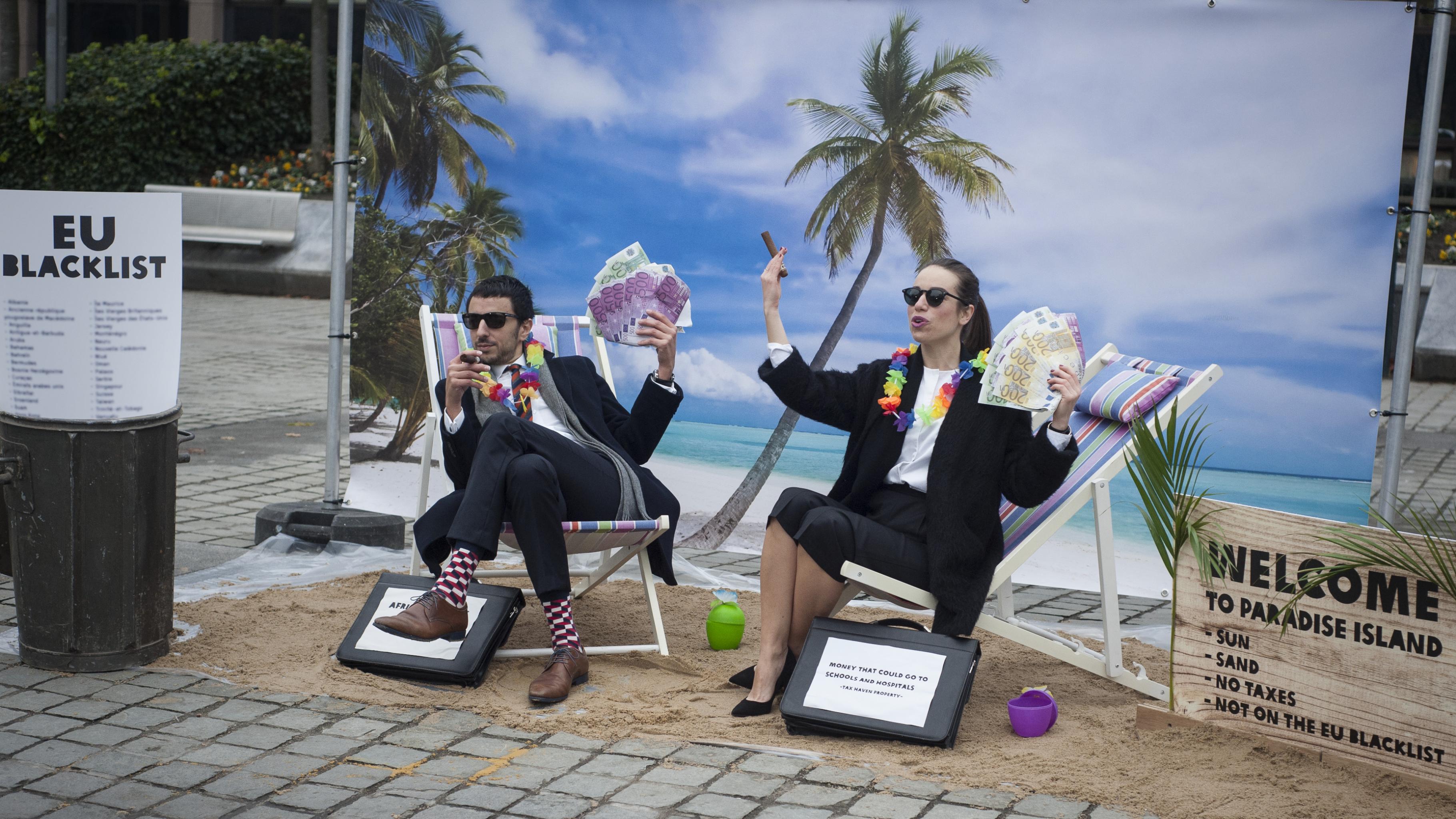 Zwei verkleidete Aktivisten im Anzug mit Sonnenbrille, Zigarren und Geldscheinen sitzen in einer karibischen Kulisse und freuen sich darüber, dass ihre Steueroase nicht auf der Schwarzen Liste der EU steht.