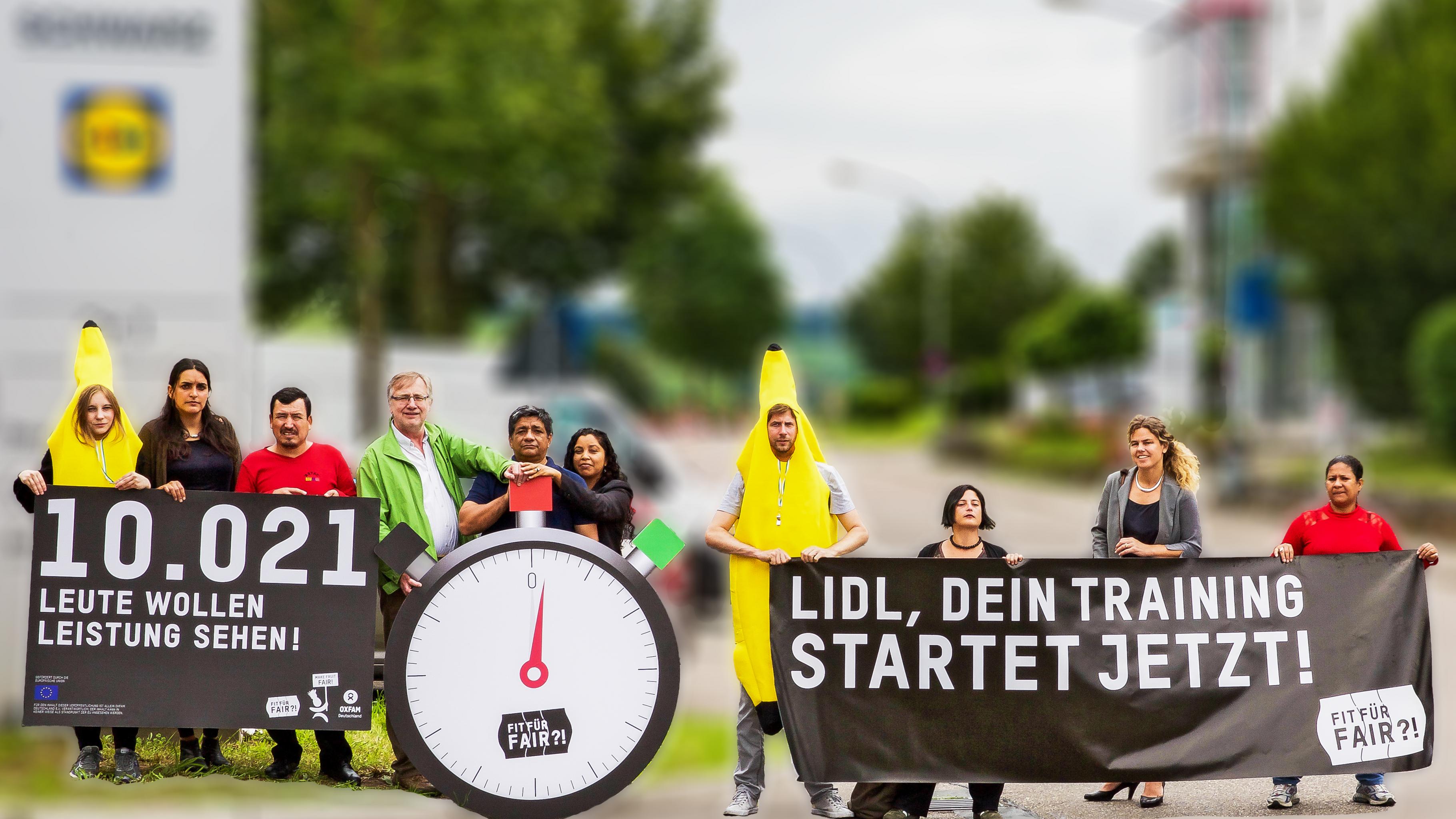 Aktivistinnen und Aktivisten, teilweise in Bananen-Kostümen, vor der Lidl-Konzernzentrale, mit Bannern „10021 Leute wollen Leistung sehen!“, „Lidl, dein Training startet jetzt!“ und einer großen Stoppuhr mit der Aufschrift „Fit für Fair?!“.