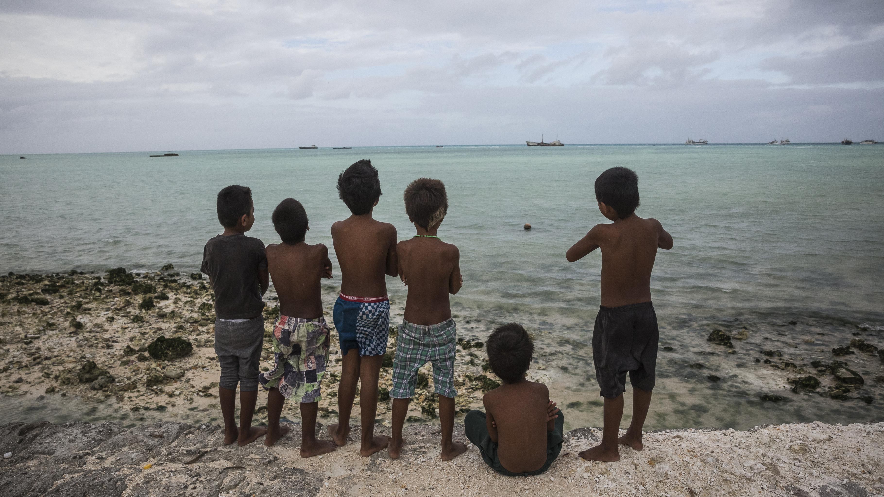 Ein Kind sitzt und fünf Kinder stehen auf einem Damm und blicken auf das Meer.