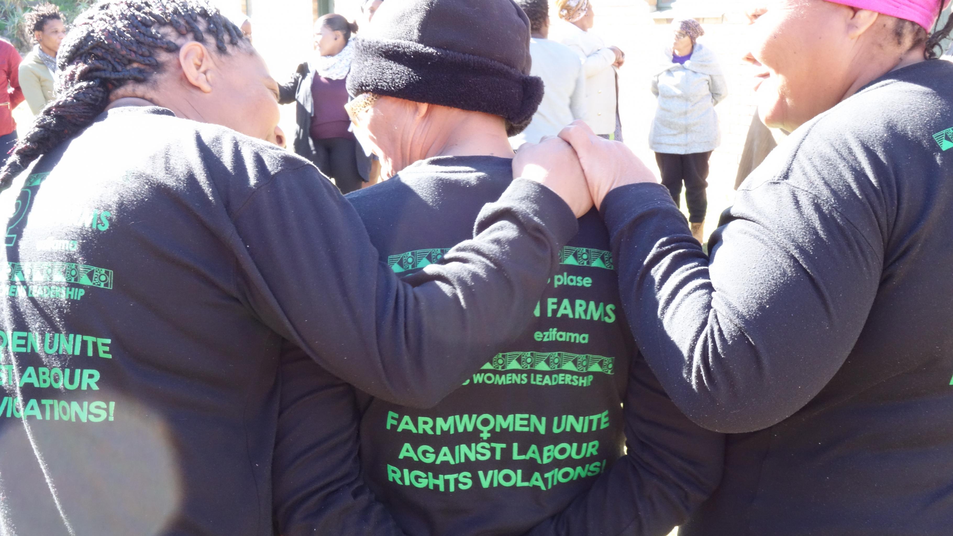 Drei Frauen stehen nebeneinander, die Hände auf die Schultern gelegt. Auf ihren Pullovern steht: „Farmwomen unite against labour rights violations!“