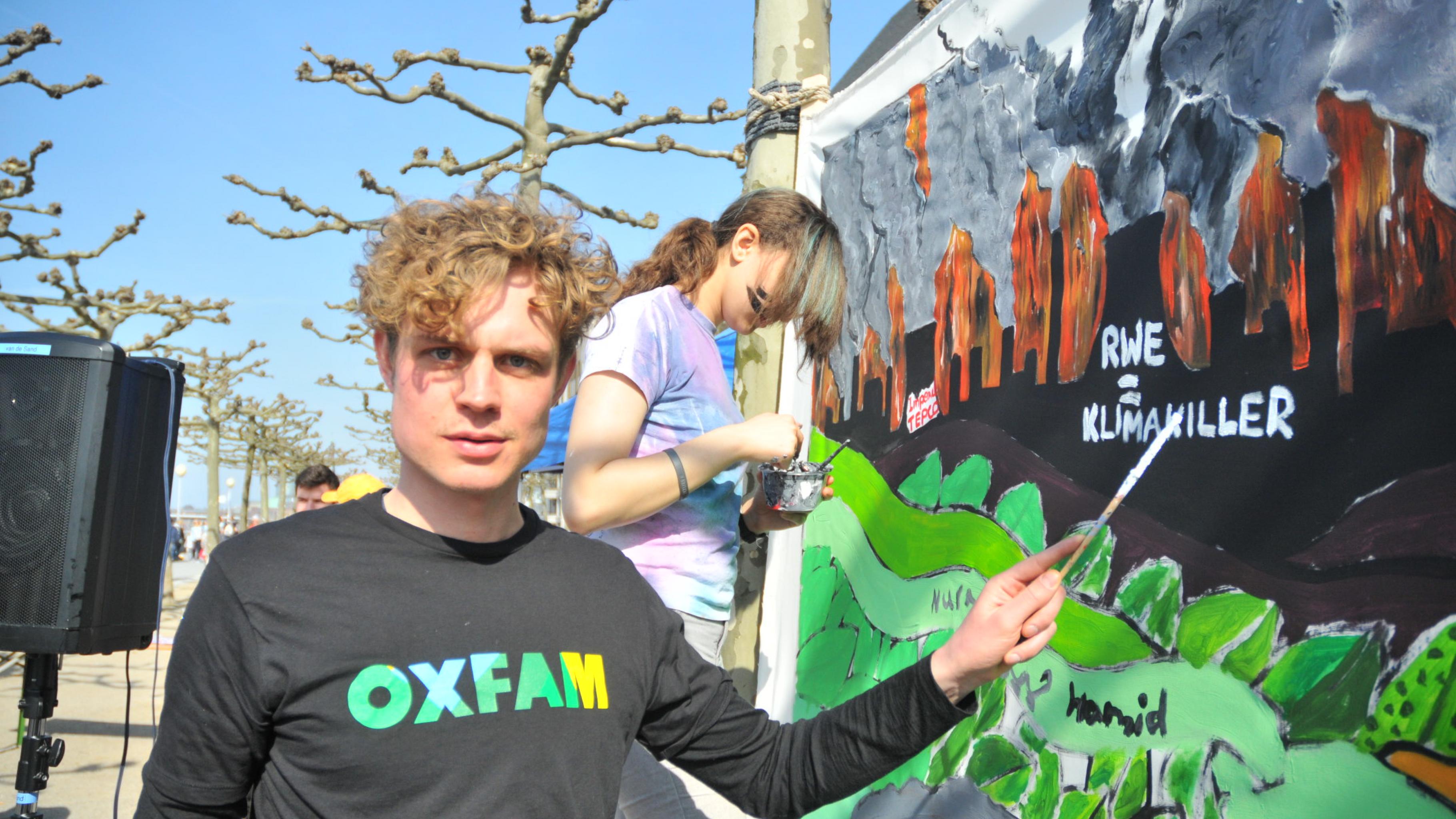 Oxfams Klima-Campaigner Bastian Neuwirth zeigt auf einen Teil des Gemäldes, Kohlekraftwerke mit dem Schriftzug „RWE = Klimakiller“ darstellt.