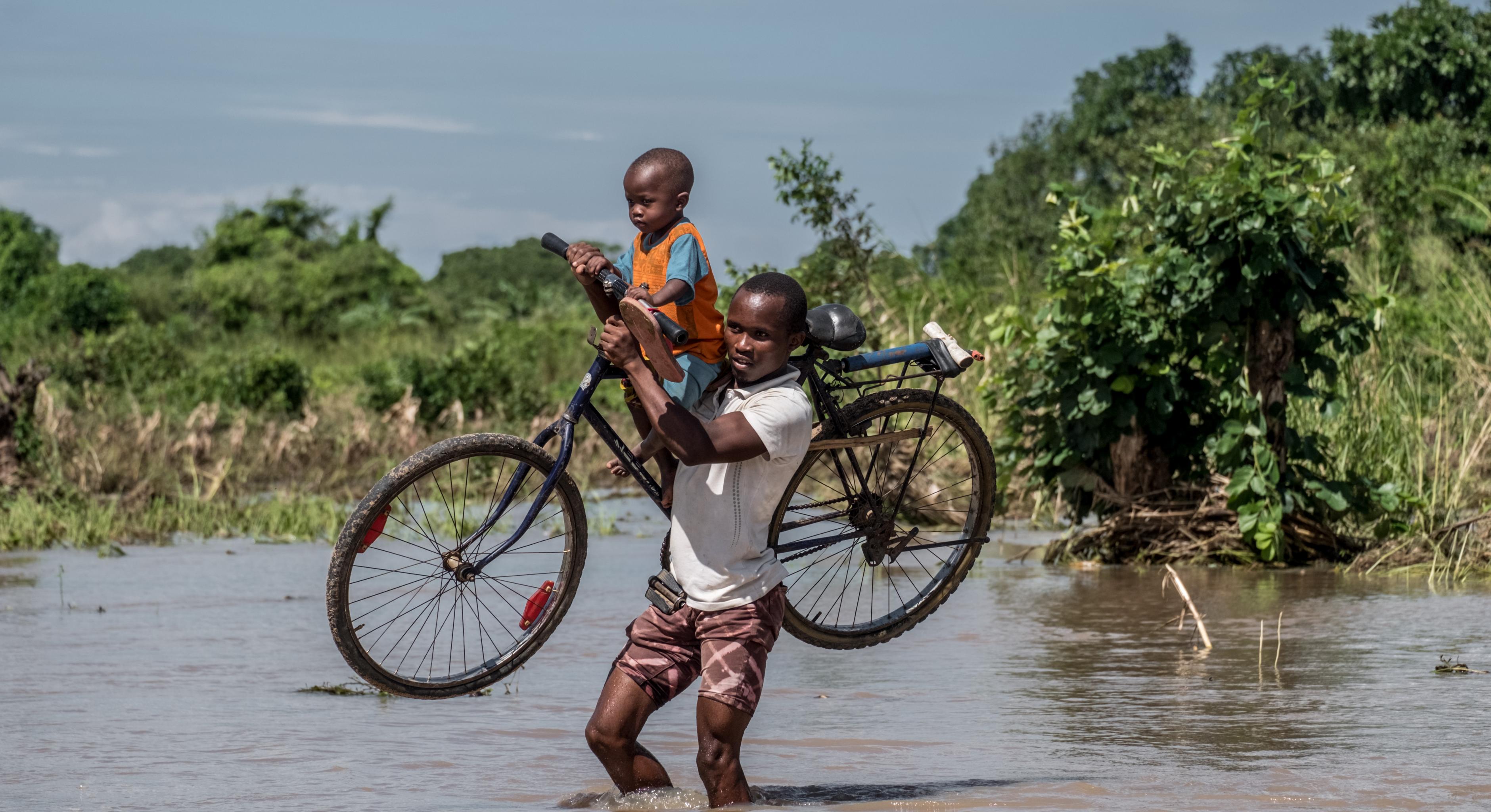 Ein Mann läuft durch Wassermassen während er ein Fahrrad und ein Kind trägt