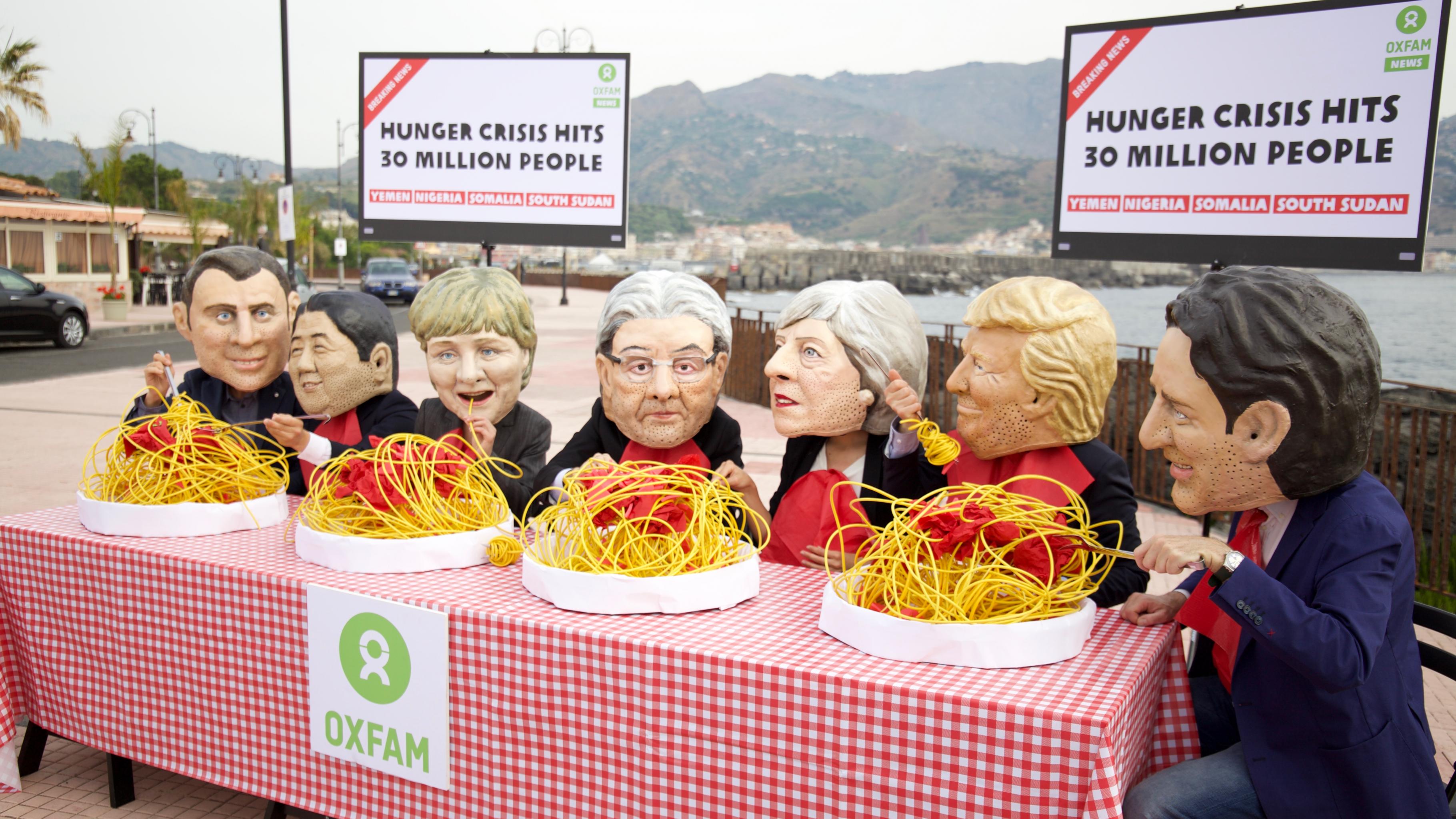 Aktivist/innen mit Masken der Staats- und Regierungschefs essen Spaghetti, während im Hintergrund auf Fernsehern die Meldung erscheint: "Hunger crisis hits 30 million people – Yemen, Nigeria, Somalia, South Sudan"