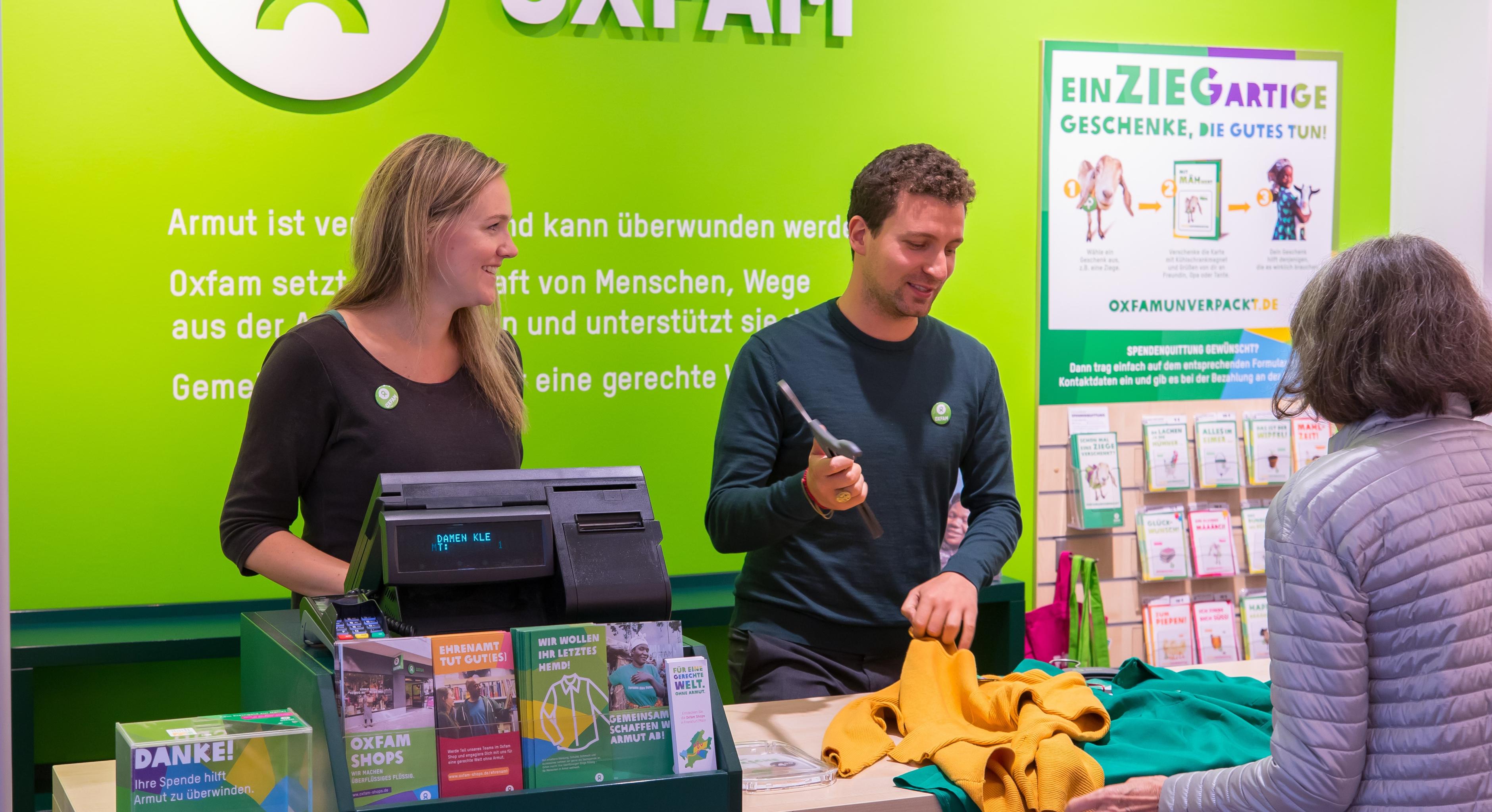 Zwei Ehrenamtliche stehen an einer Kasse in einem Oxfam-Shop und kassieren die Kleidung einer Kundin ab