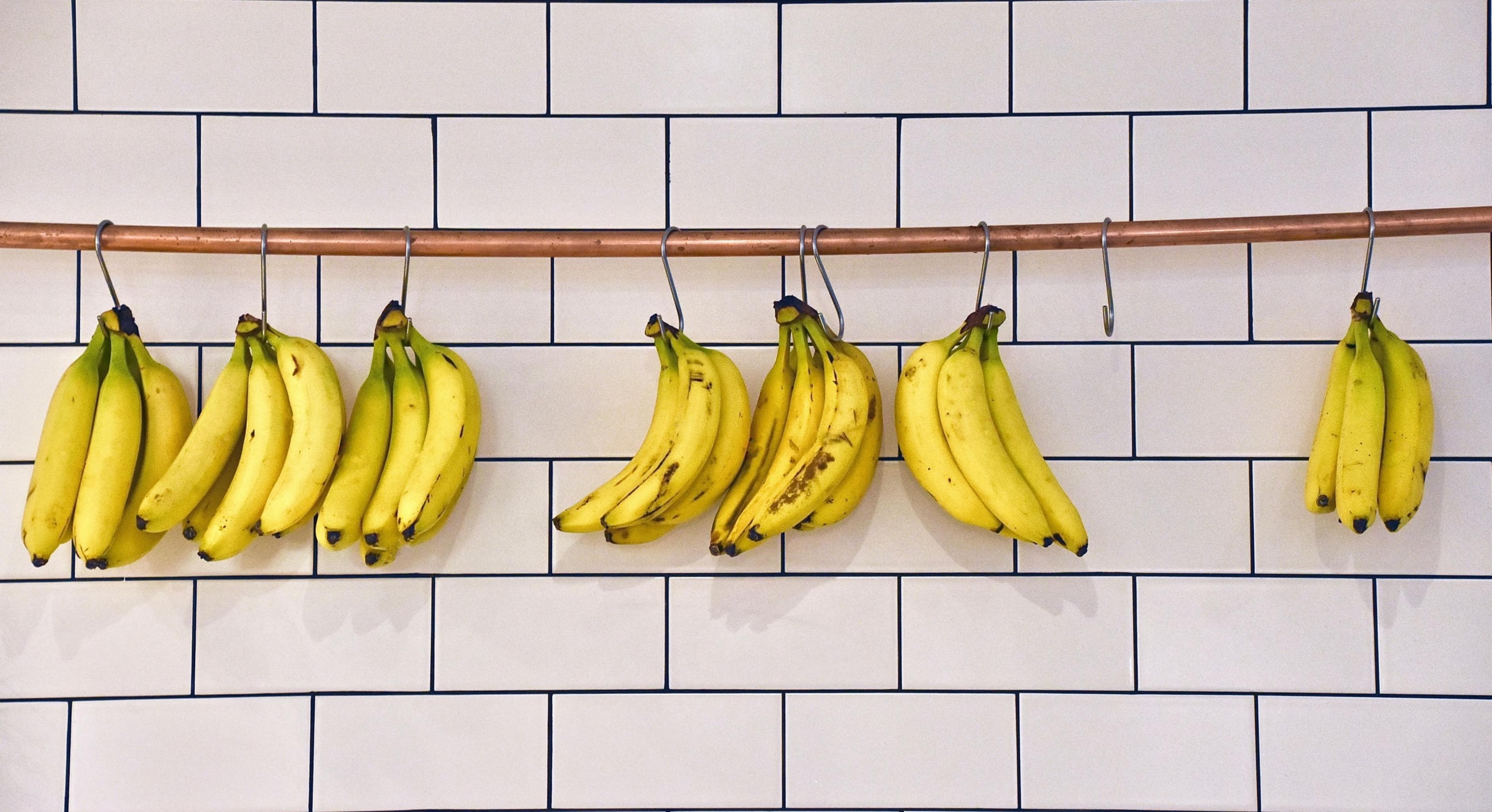 Bananen hängen an einer Stange vor einer weiß gefliesten Wand.