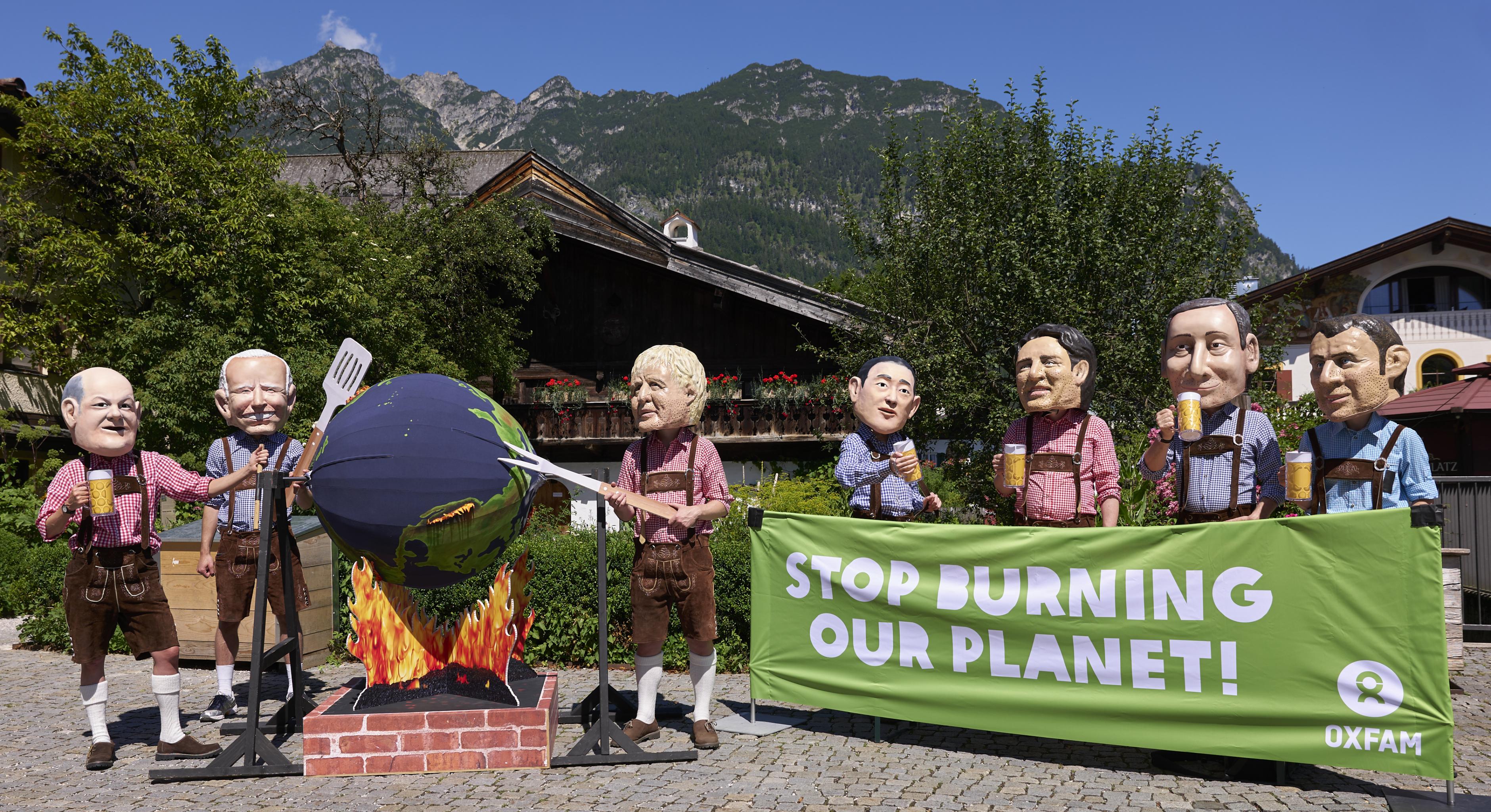 In Lederhosen und vor alpiner Kulisse grillen die Bigheads (Karikaturen der Staats- und Regierungschefs der G7) die Erde. Oxfam fordert: „Stop burning our planet!“