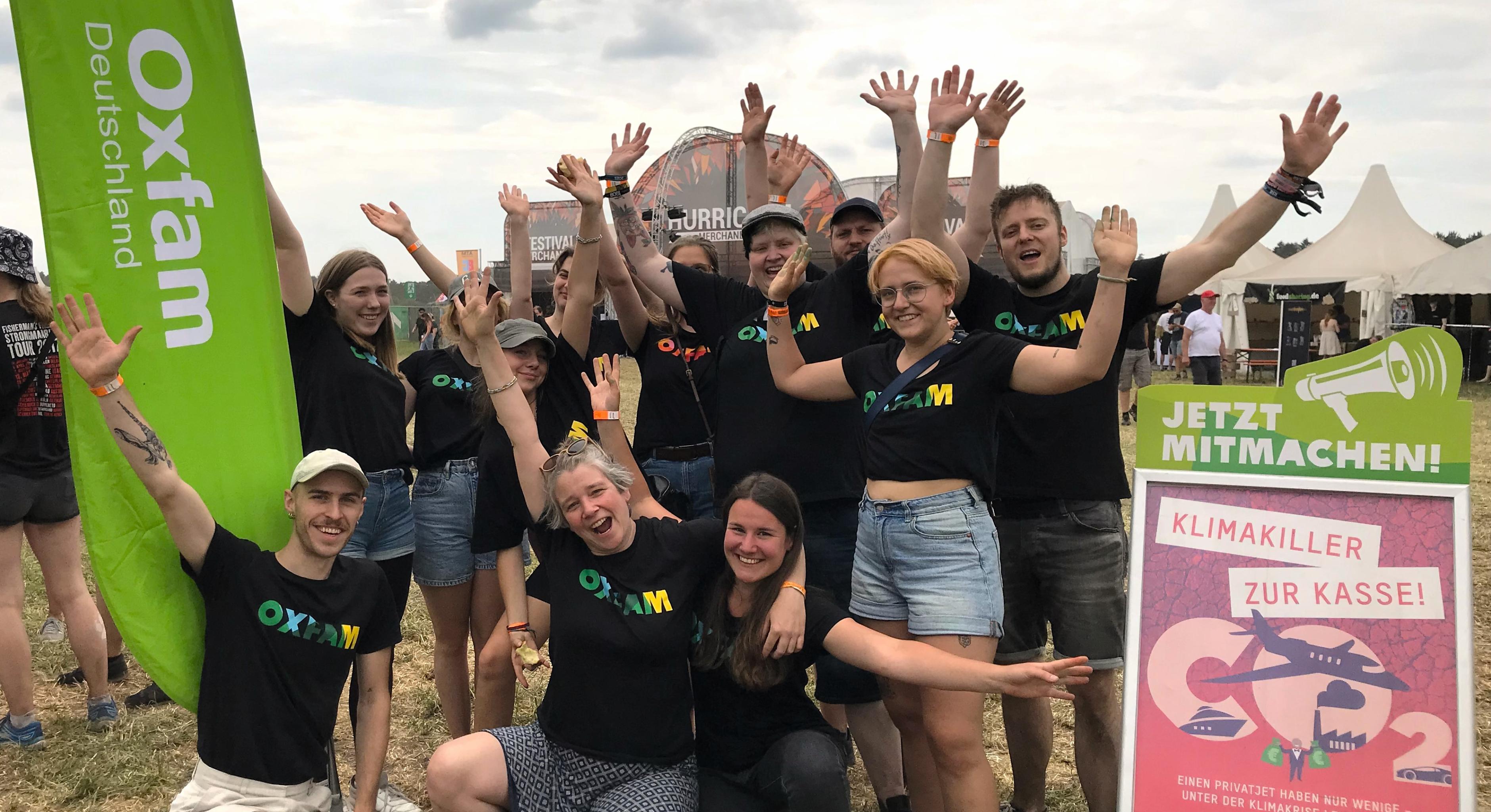 Gruppenfoto von Aktivist*innen in Oxfam-Shirts, sie strecken die Arme in die Luft und lächeln. Neben ihnen befinden sich ein grüner Oxfam-Banner und ein Info-Schild zur aktuellen Kampagne. Im Hintergrund ist das Hurricane-Festivalgelände zu sehen.