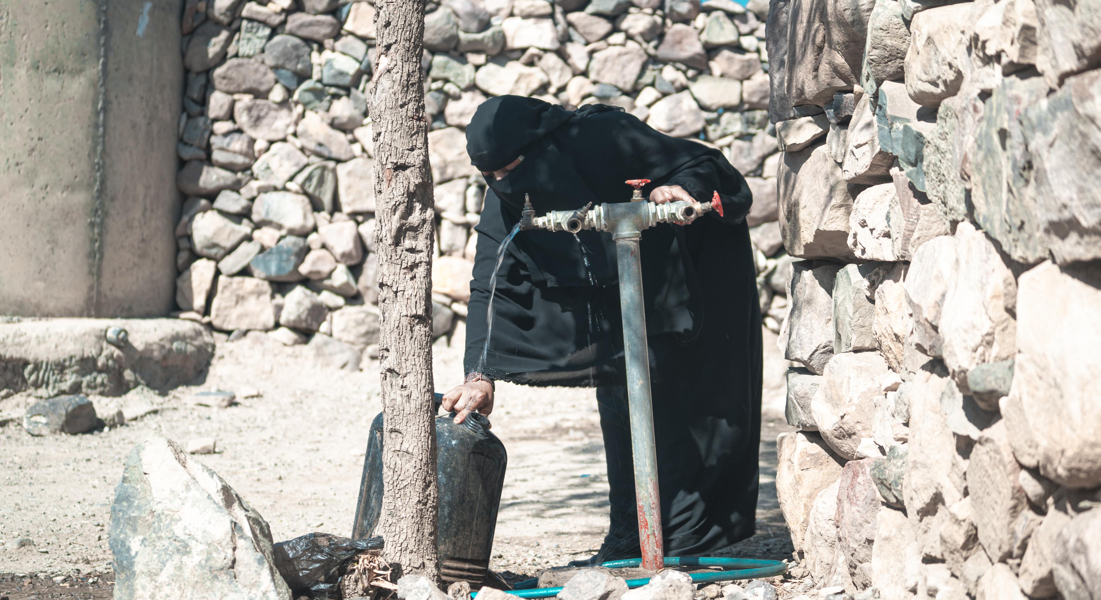 Haneen* (in schwarzem Gewand und mit Niqab bekleidet) dreht einen Wasserhahn vor ihrem Haus auf, um einen Wasserkanister zu befüllen.