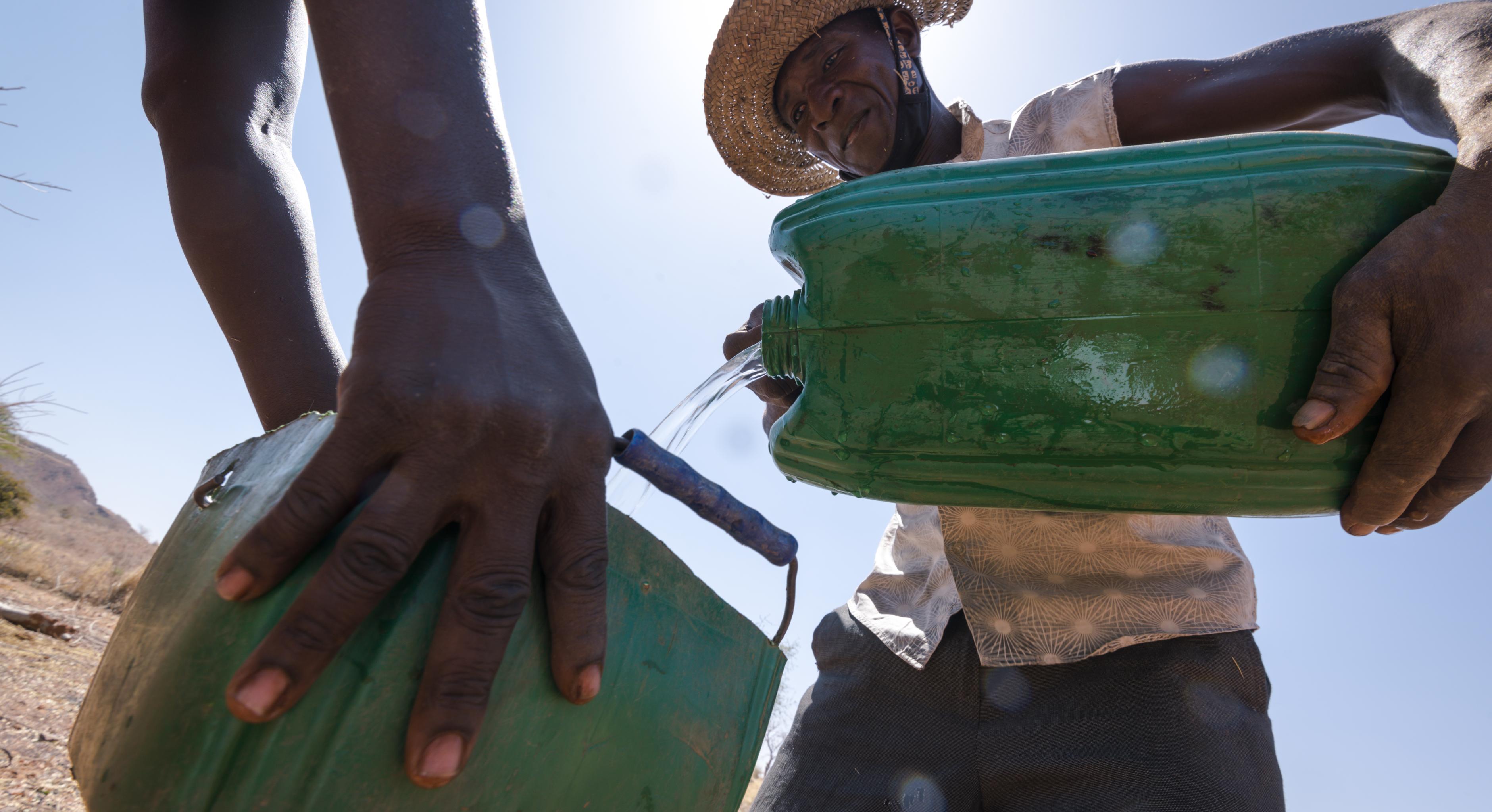 Samuel Sawadogo teilt Wasser auf zwei grüne Wasserkanister auf. Er trägt einen Hut zum Schutz vor der Sonne, ein beige-gemustertes Hemd und eine dunkle Hose. Man sieht links die Hände einer weiteren Person, die den zweiten Kanister hält.