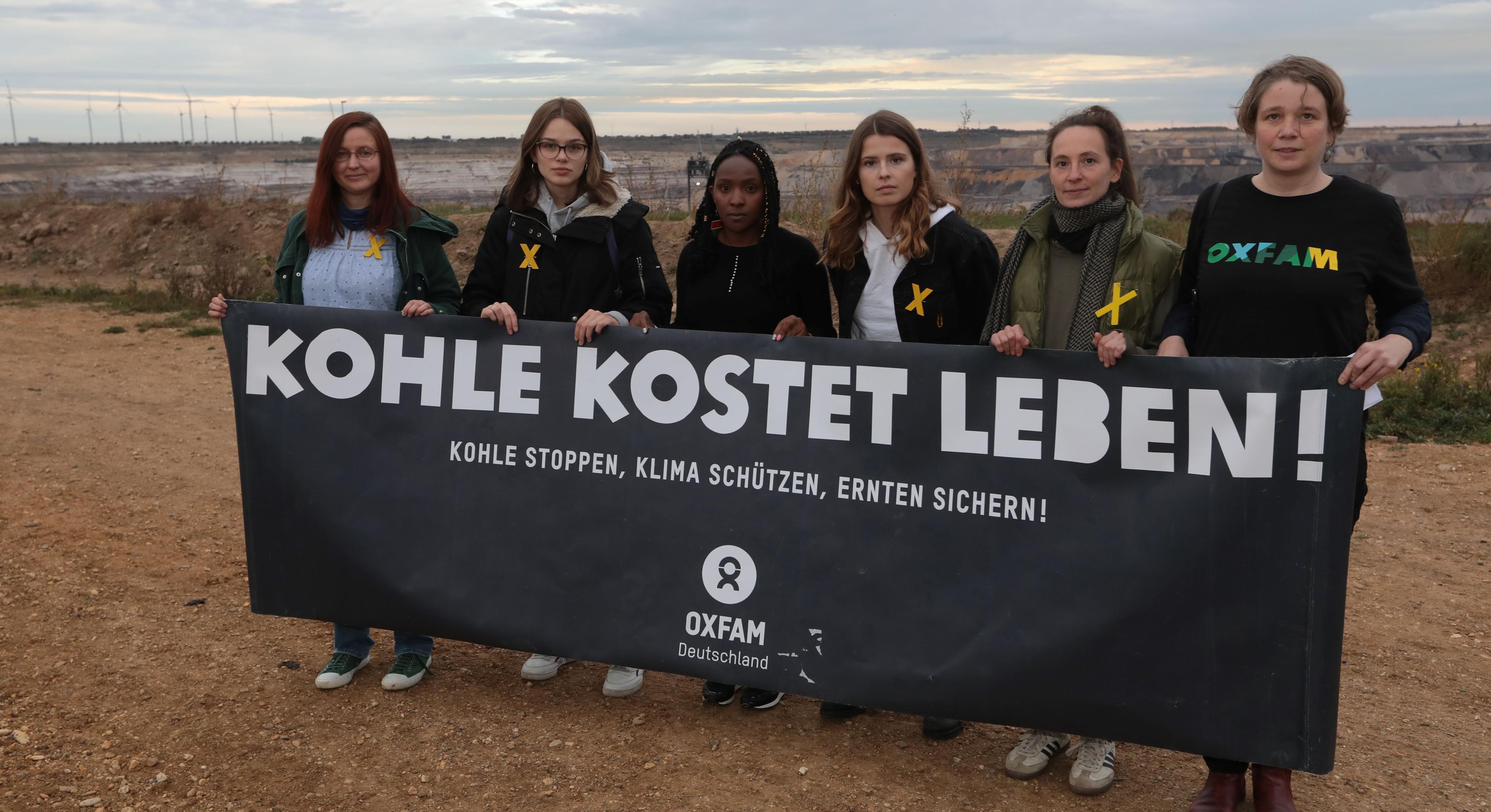 Sechs Frauen stehen mit einem schwarzen Banner in der Hand am Rand des Tagebaus Garzweiler. Auf dem Banner steht: Kohle kostet leben! Kohle stoppen, Klima schützen, Ernten sichern!