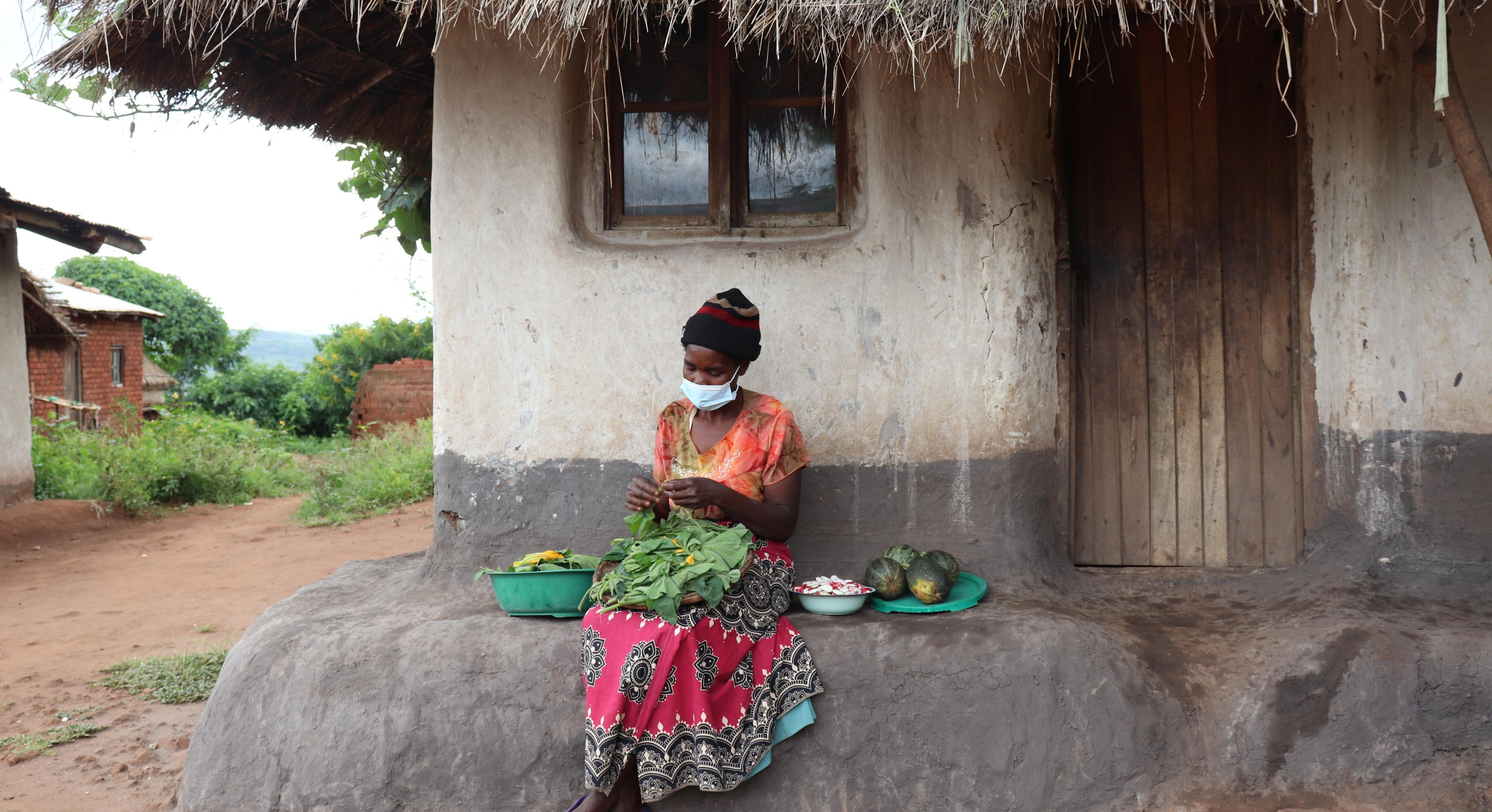 Loveness N'Dindo. mit einem rot-schwarz gemusterten Rock und orangenen T-Shirt sowie Mundschutz bekleidet, sitzt auf einer Steinbank vor ihrem Haus. Auf ihrem Schoß sortiert sie Gemüse. Neben ihr befinden sich weitere Schüsseln mit Früchten. 