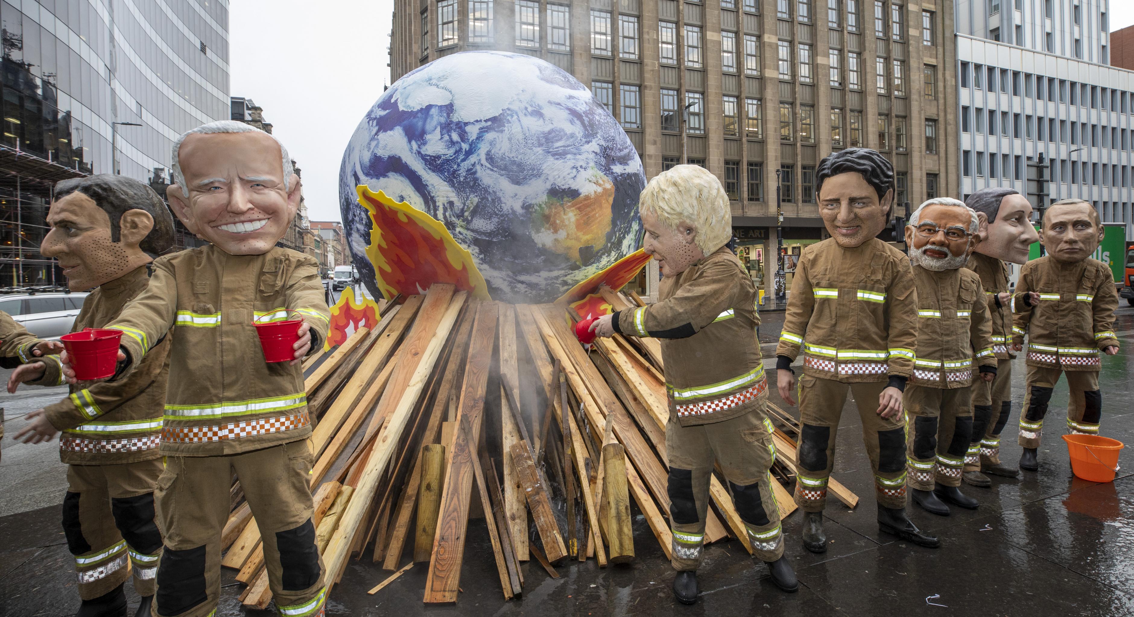 Oxfam-Aktivist*innen posieren mit überdemensionalen Pappmasche-Köpfen der mächtigsten Regierungspersonen und als Feuerwehrmenschen verkleidet bei der COP26 in Glasgow. Sie stehen vor einer brennenden Erdkugel und versuchen sie erfolglos zu löschen.