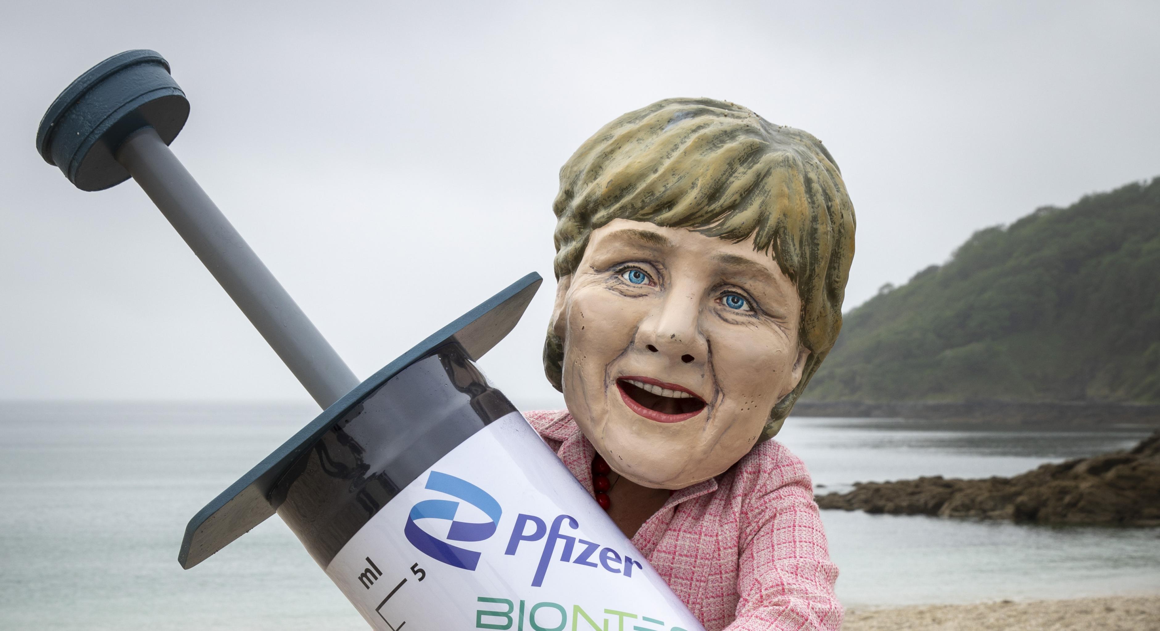 Ein*e Aktivist*in ist mit einem riesigen Angela-Merkel-Kopf bekleidet hält eine riesige Spritzen-Atrappe im Arm. Im Hintergrund sind Strand und Meer zu sehen.