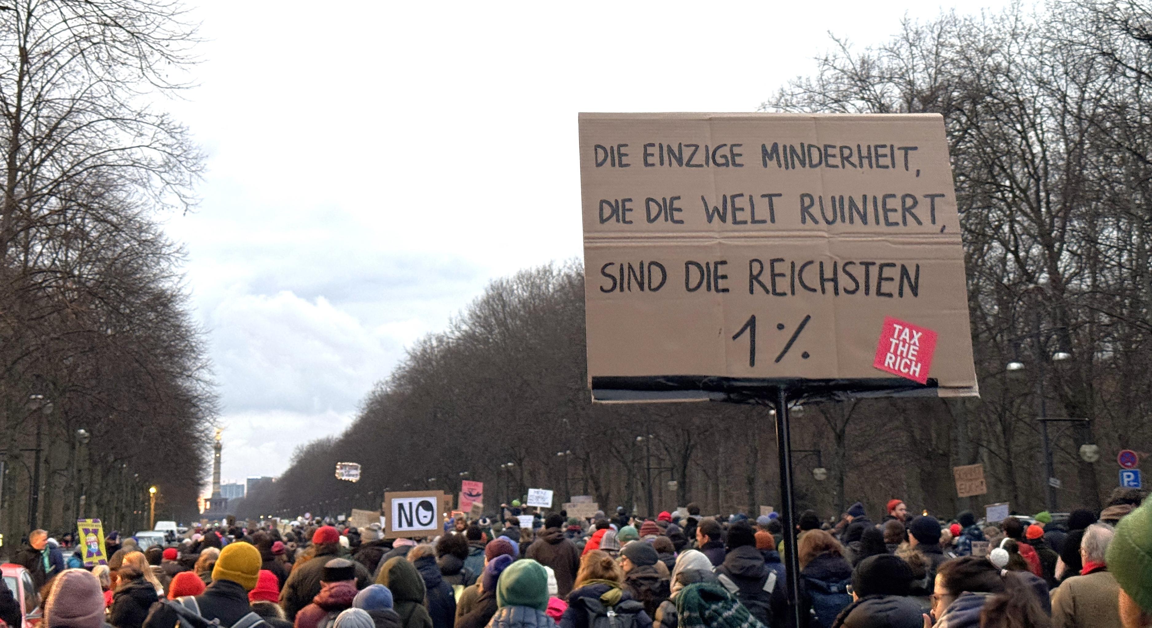 viele Menschen mit Demoschildern bewegen sich auf den Großen Stern in Berlin zu. Auf einem Schild steht "Die einzige Minderheit, die die Welt ruiniert, sind die reichsten 1 %"