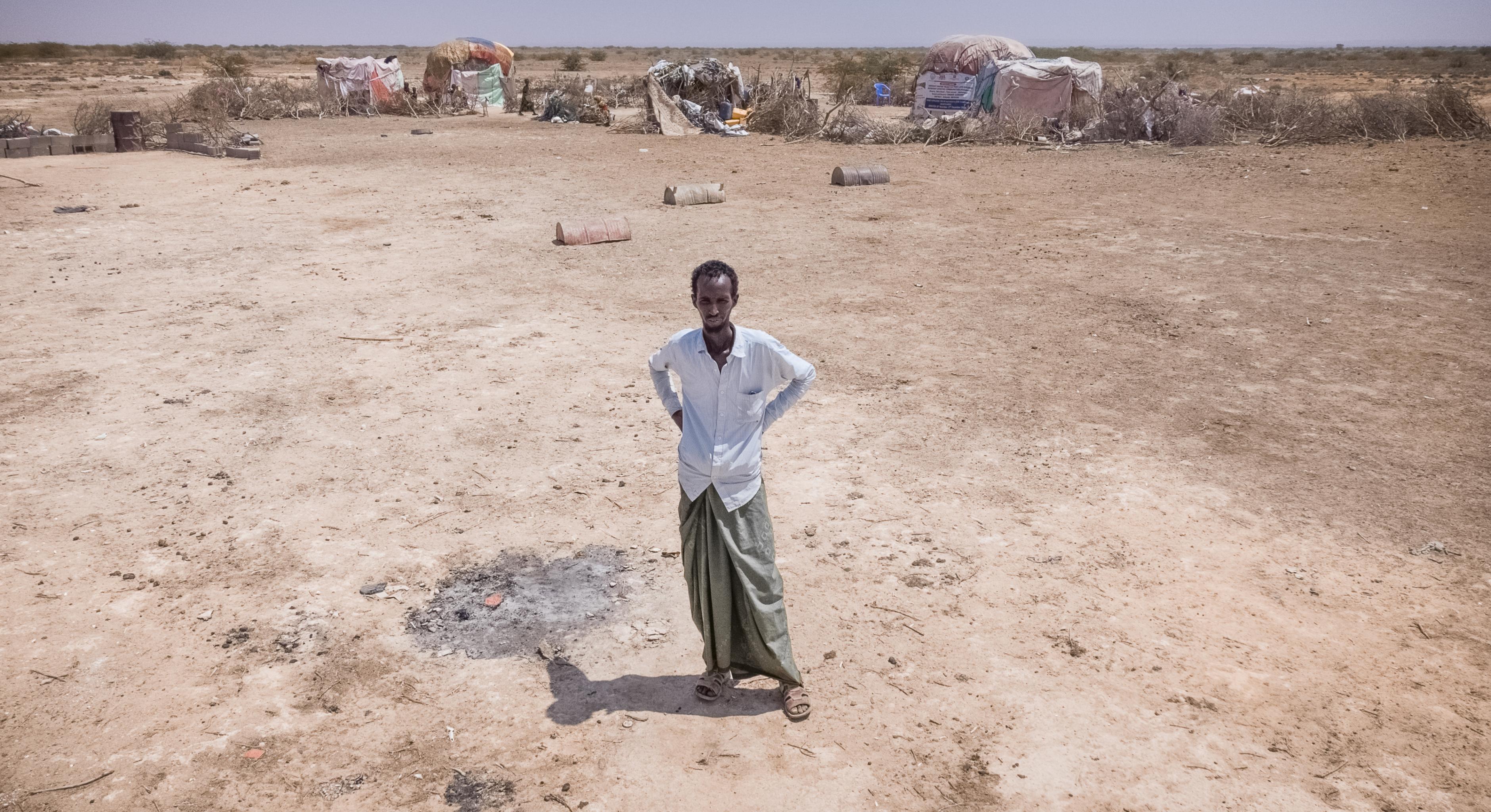 Abdulahi Farah Isse steht, bekleidet mit einem weißen Shirt und einem grauen Wickelrock, auf einem sehr trockenen Boden. Im Hintergrund ist seine Unterkunft zu sehen.