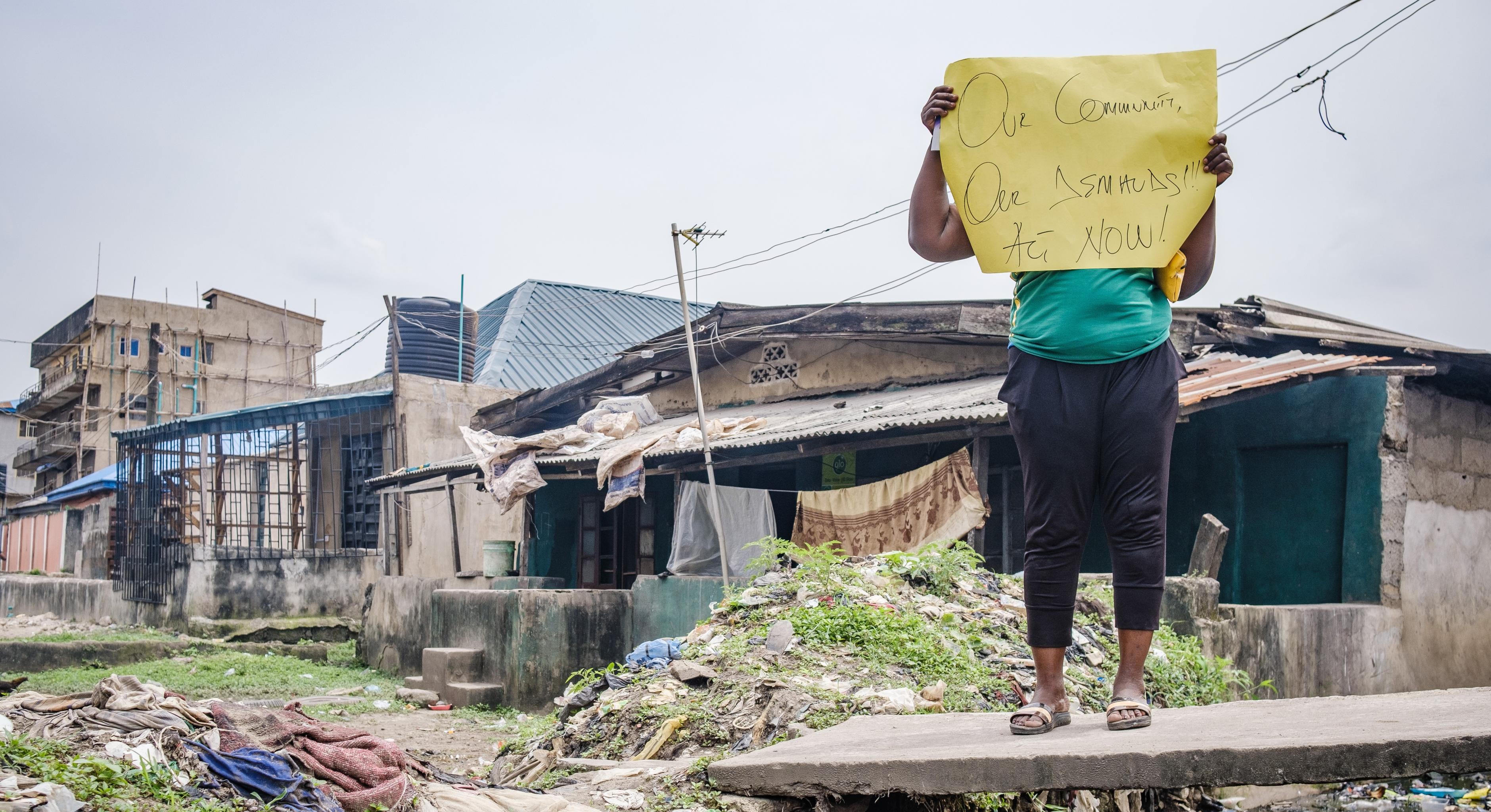Eine Aktivistin hält ein Plakat mit der Aufschrift "Unsere Community, unsere Forderungen! Jetzt handeln!" hoch. Sie trägt ein grünes T-Shirt und eine dunkle Hose. Im Hintergrund sind Häuser zu sehen.