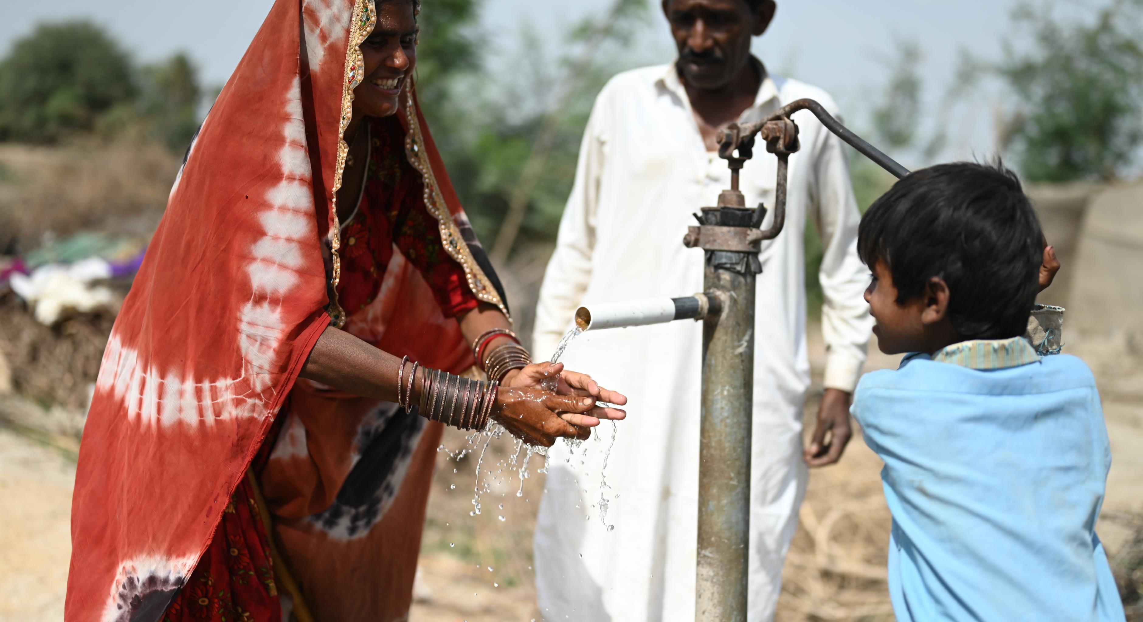 Eine Familie benutzt eine neu installierte Hand-Wasserpumpe. Das Kind (rechts) bedient die Pumpe, während die Mutter (links) ihre Hände unter den Wasserstrahl hält. Im Hintergrund steht eine dritte Person.