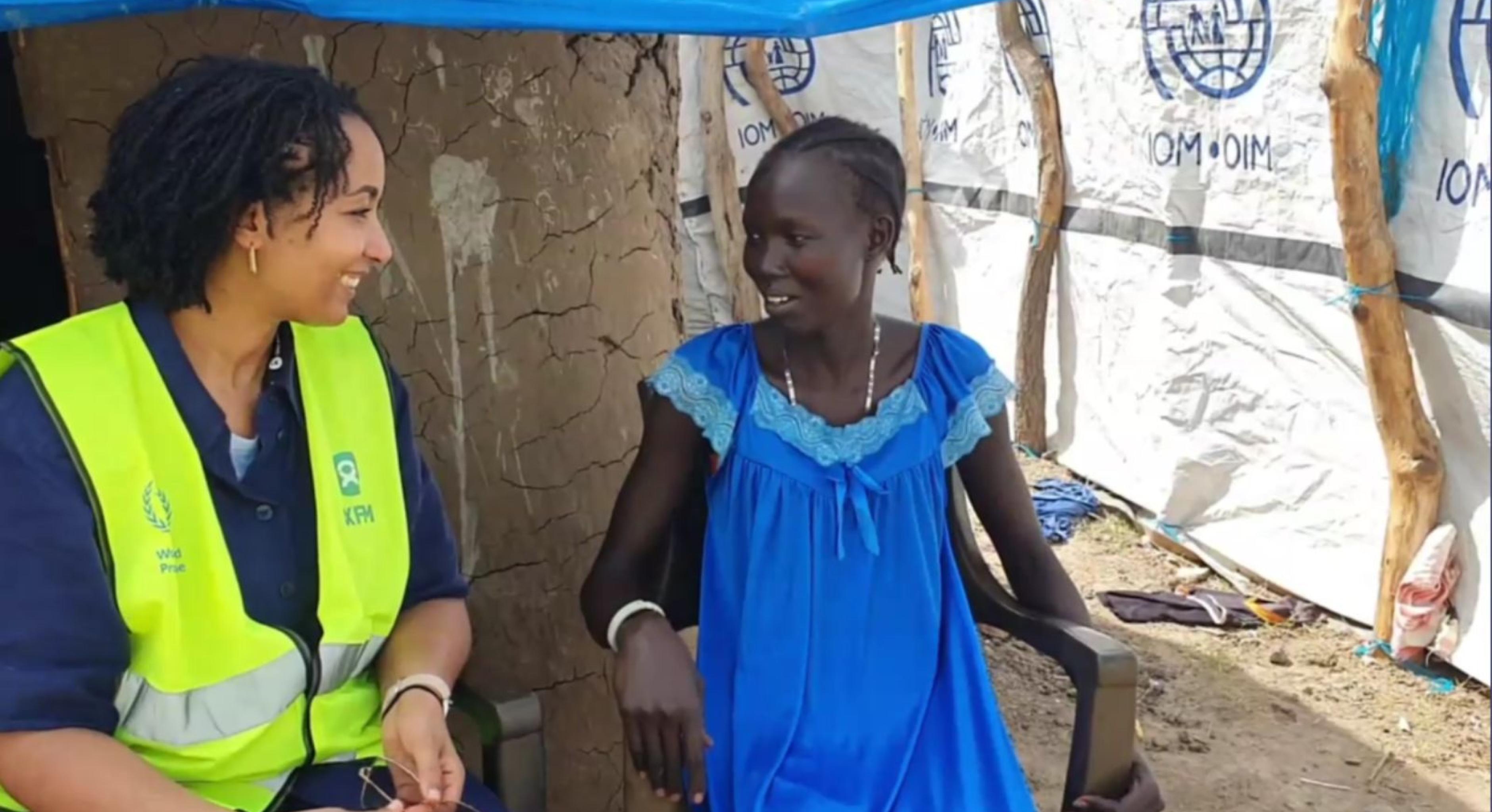 Die Direktorin von Oxfam in Afrika, Fati N'zi-Hassane (links, trägt ein dunkelblaues Hemd und eine neongelbe Weste) und Nyaguka Jiek (rechts, trägt ein blaues Kleid) sitzen auf weißen Plastikstühlen nebeneinander und unterhalten sich. 