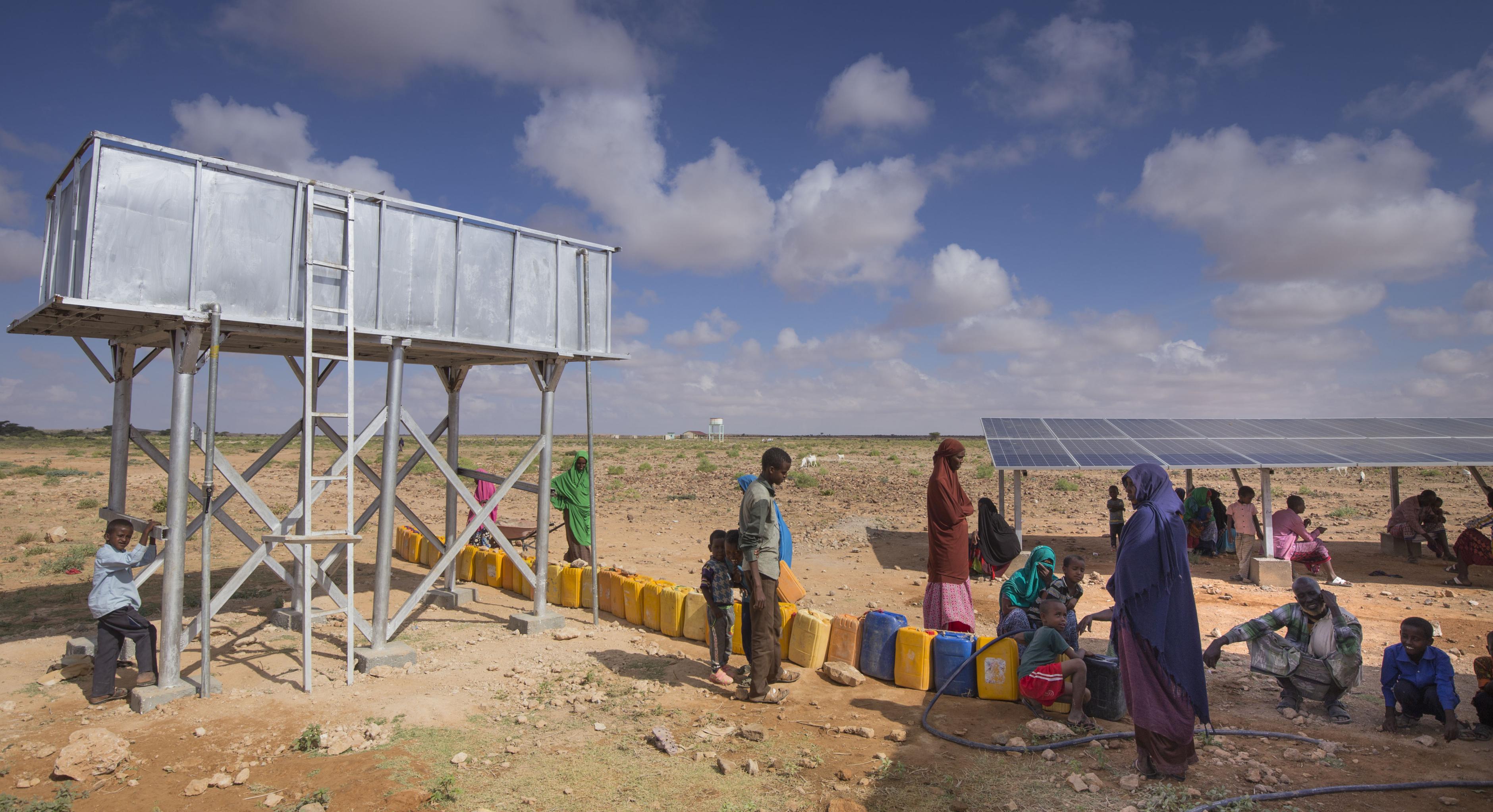 In der von Oxfam gebauten Entsalzungsanlage in Ceel Midgaan, Somaliland, wird sauberes Wasser erzeugt.Das Bild zeigt einen Wassertank, vor dem viele Wasserkanister stehen. Im Hintergrund steht eine Solaranlage, die die Entsalzungsanlage mit Strom versorgt. Unter den Solarpanels sitzen einige Menschen im Schatten. Weitere Menschen stehen und Hocken in der Sonne und warten darauf, ihre Kanister befüllen zu können.