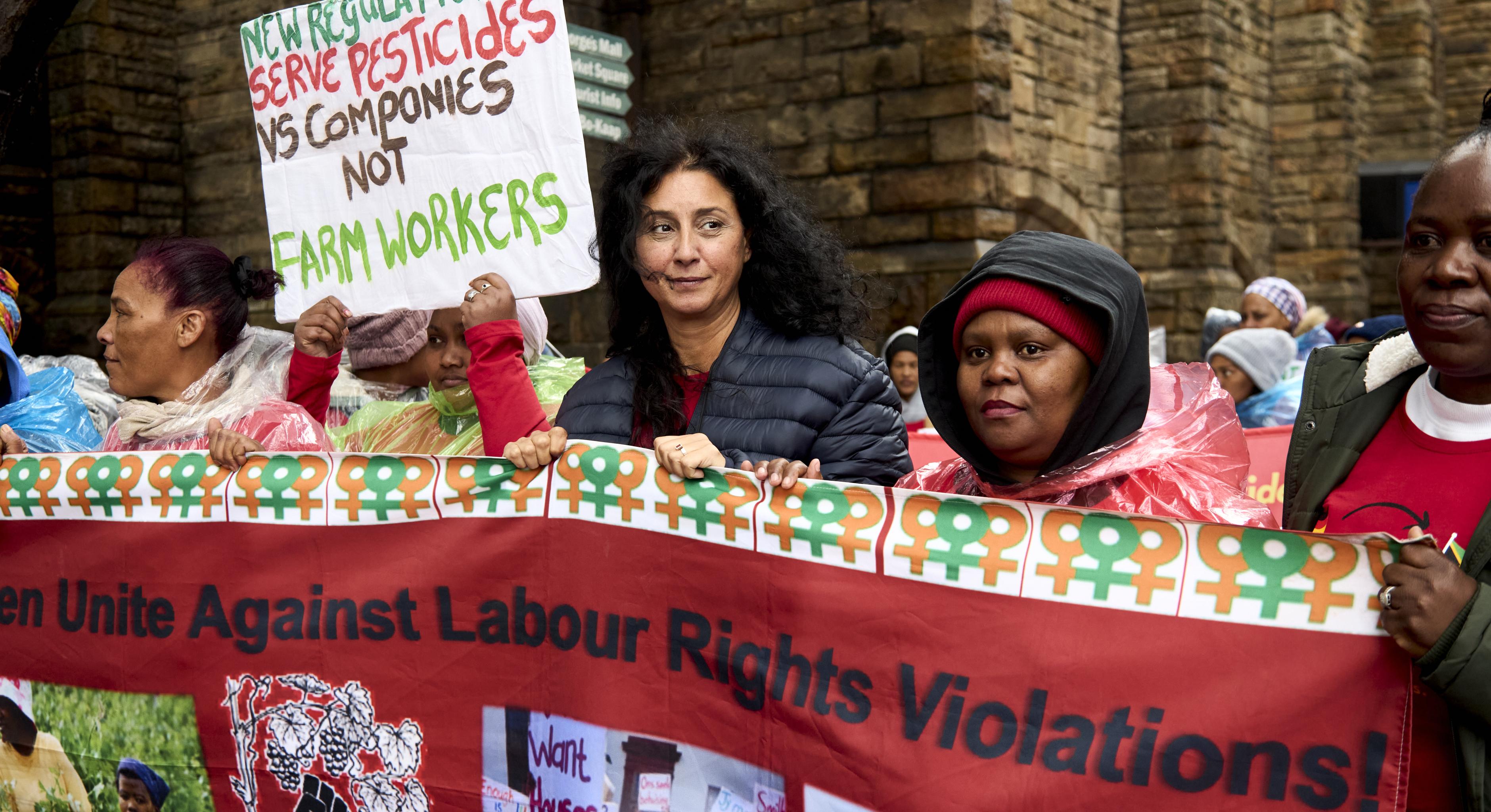 Frauen auf einer Demonstration, zu lesen ist „Unite against labor rights violations“ und „New regulations serve pesticides vs companies not farm workers“