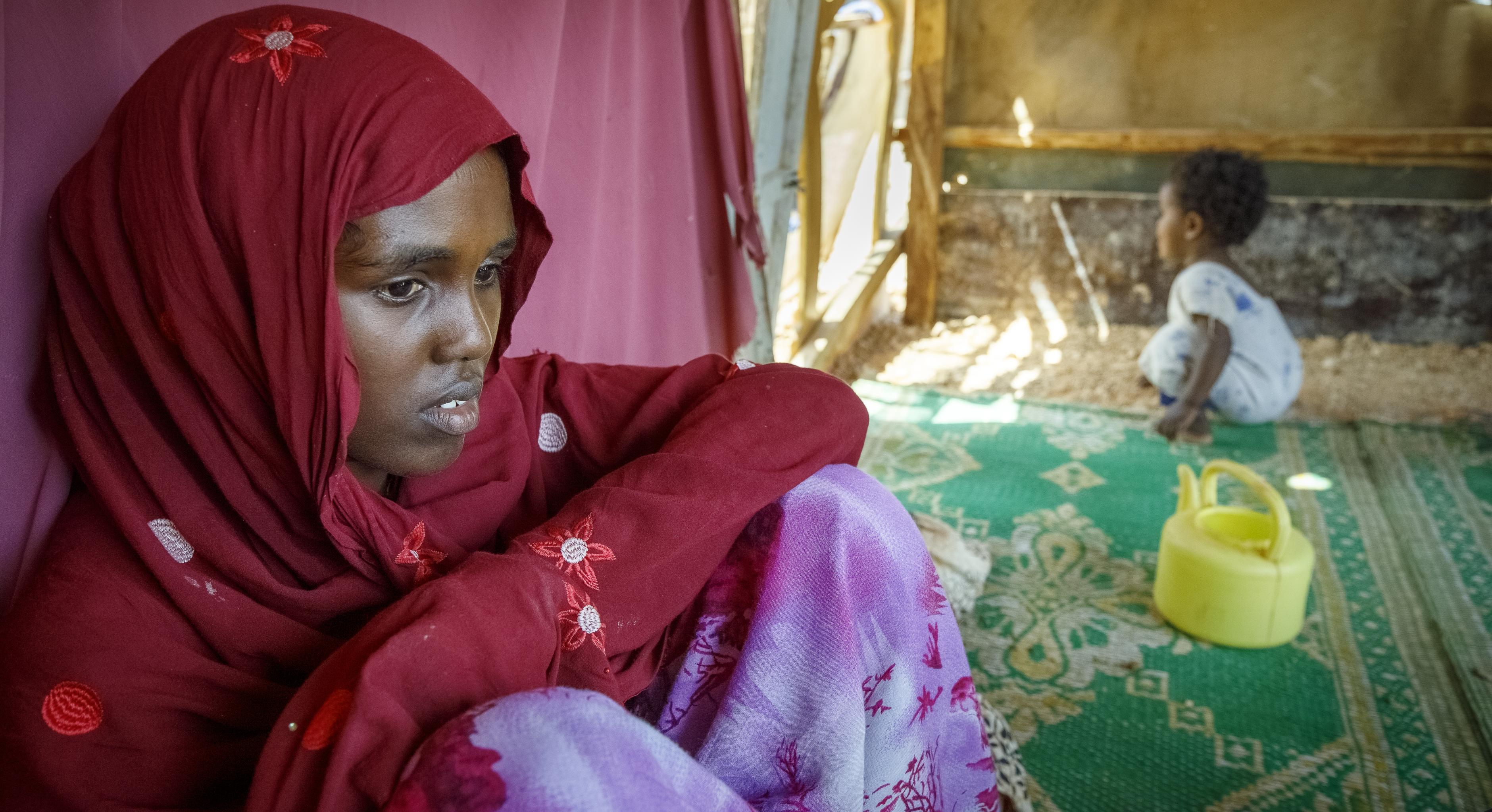 Hafsa Abdikader Ahmed, bekleidet mit einem roten Oberteil und Kopftuch sowie einer lila Hose, sitzt mit herangezogenen Knien auf einem grünen Teppich. Im Hintergrund läuft ein Kind an ihr vorbei. 