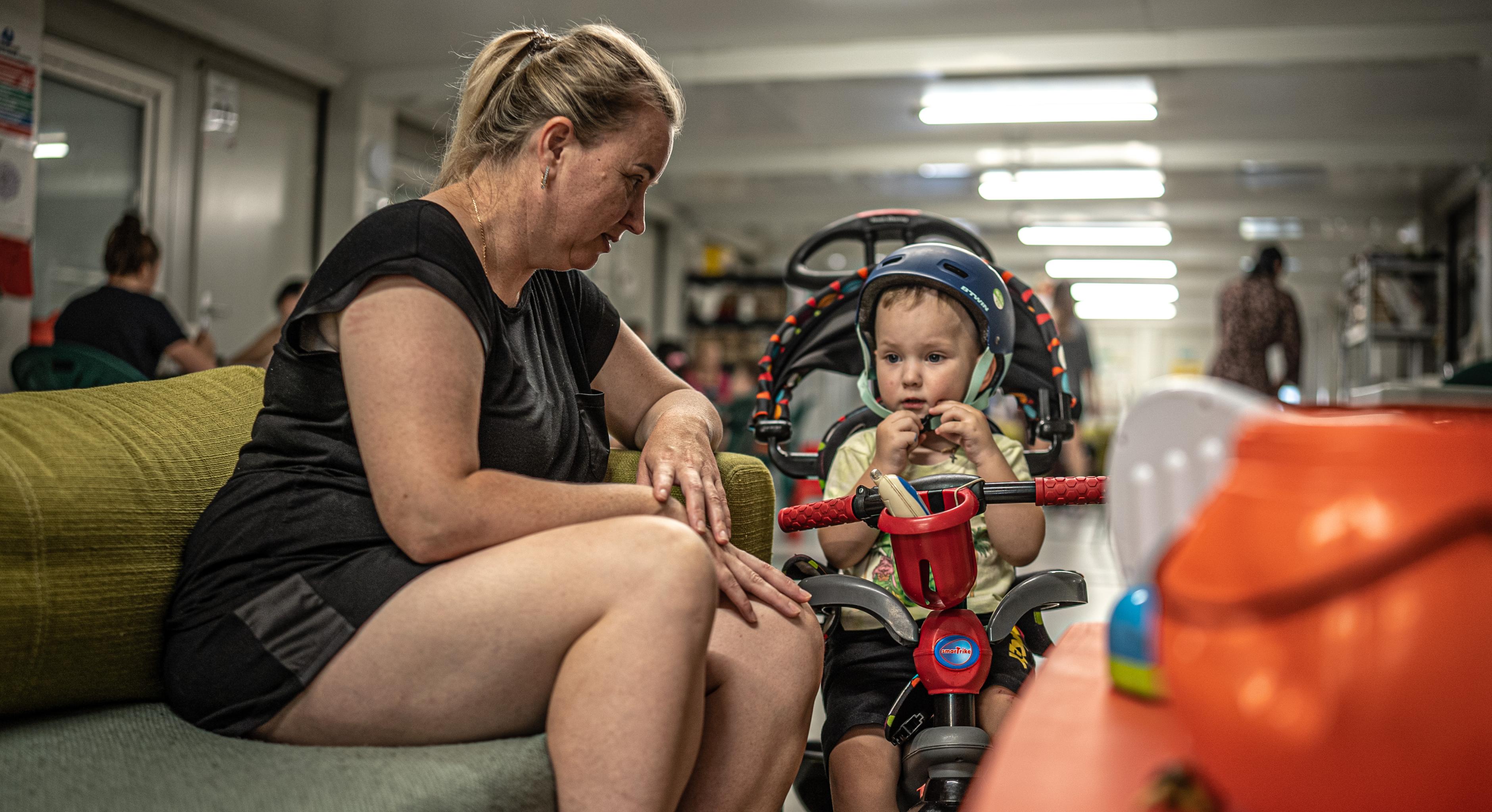 Eine Frau sitzt in einem großen Raum auf einem Sofa. Ihr Kopf ist dem Kinderwagen zugeneigt, der neben ihr steht und in dem ihr Sohn sitzt.