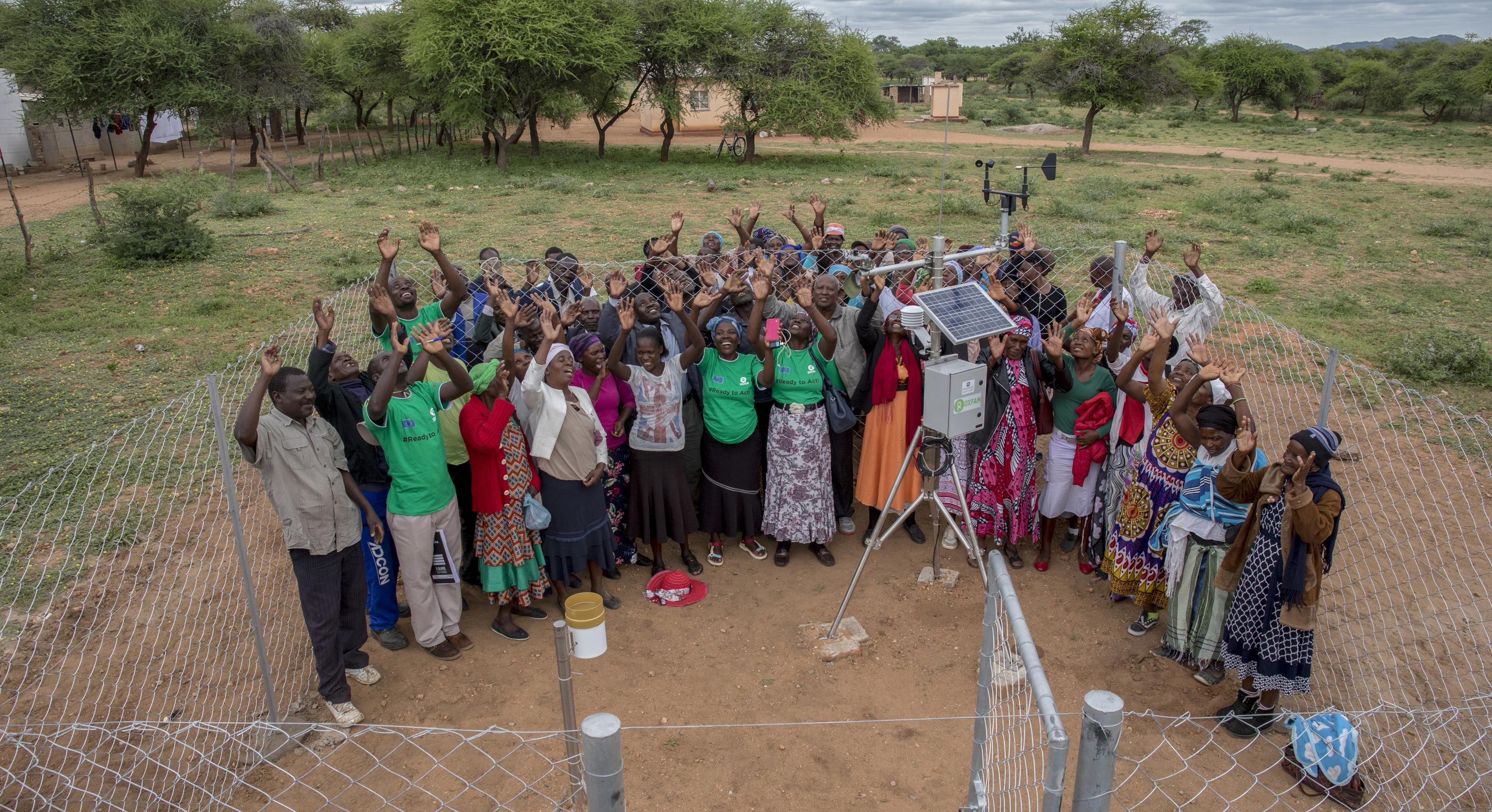 Eine Gruppe von Menschen steht bei einer Wetterstation in einer ländlichen Region in Simbabwe. Sie strecken freudig ihre Arme in die Luft.