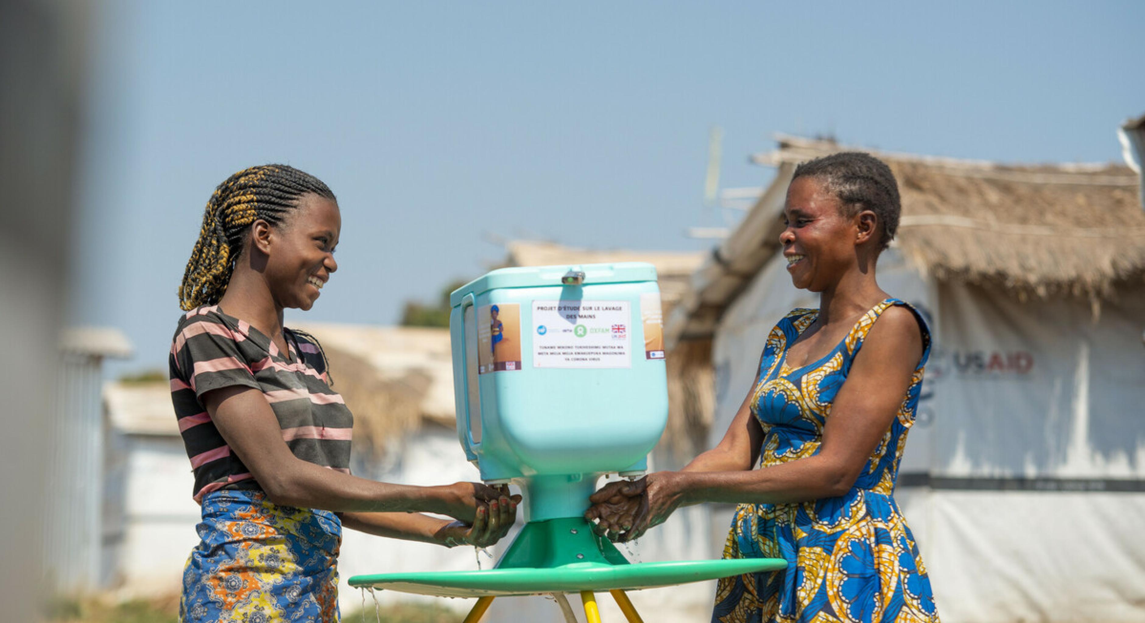 Zwei Frauen waschen ihre Hände an einer Handwasch-Station im Geflüchtetencamp in Kisalaba (Demokratische Republik Kongo). Die Frauen schauen einander lächelnd an.