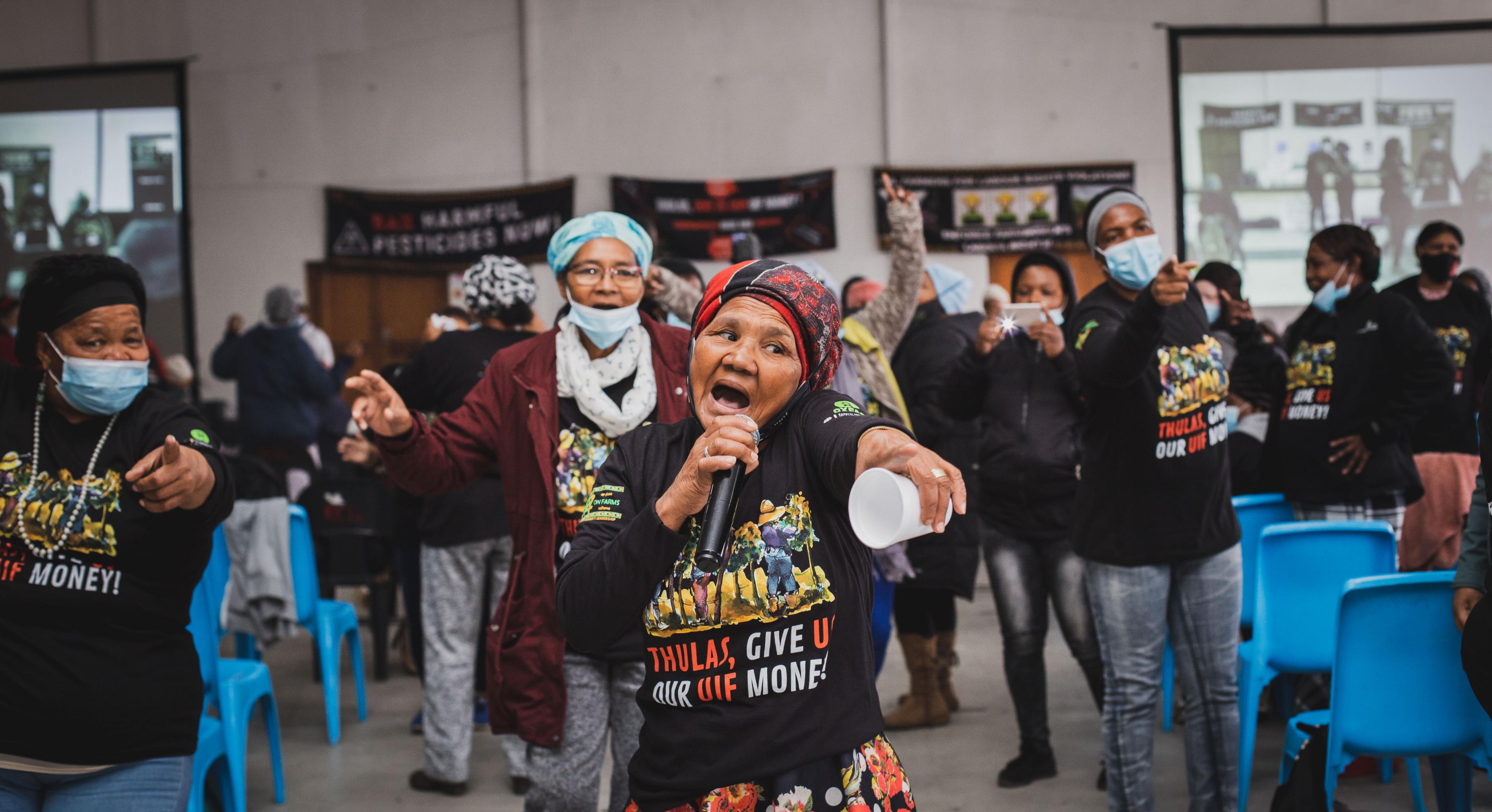 Eine Gruppe von Farmarbeiterinnen singen auf einer Protestaktion mit schwarzen Tshirts mit der Aufschrift "Thulas, give us our money".