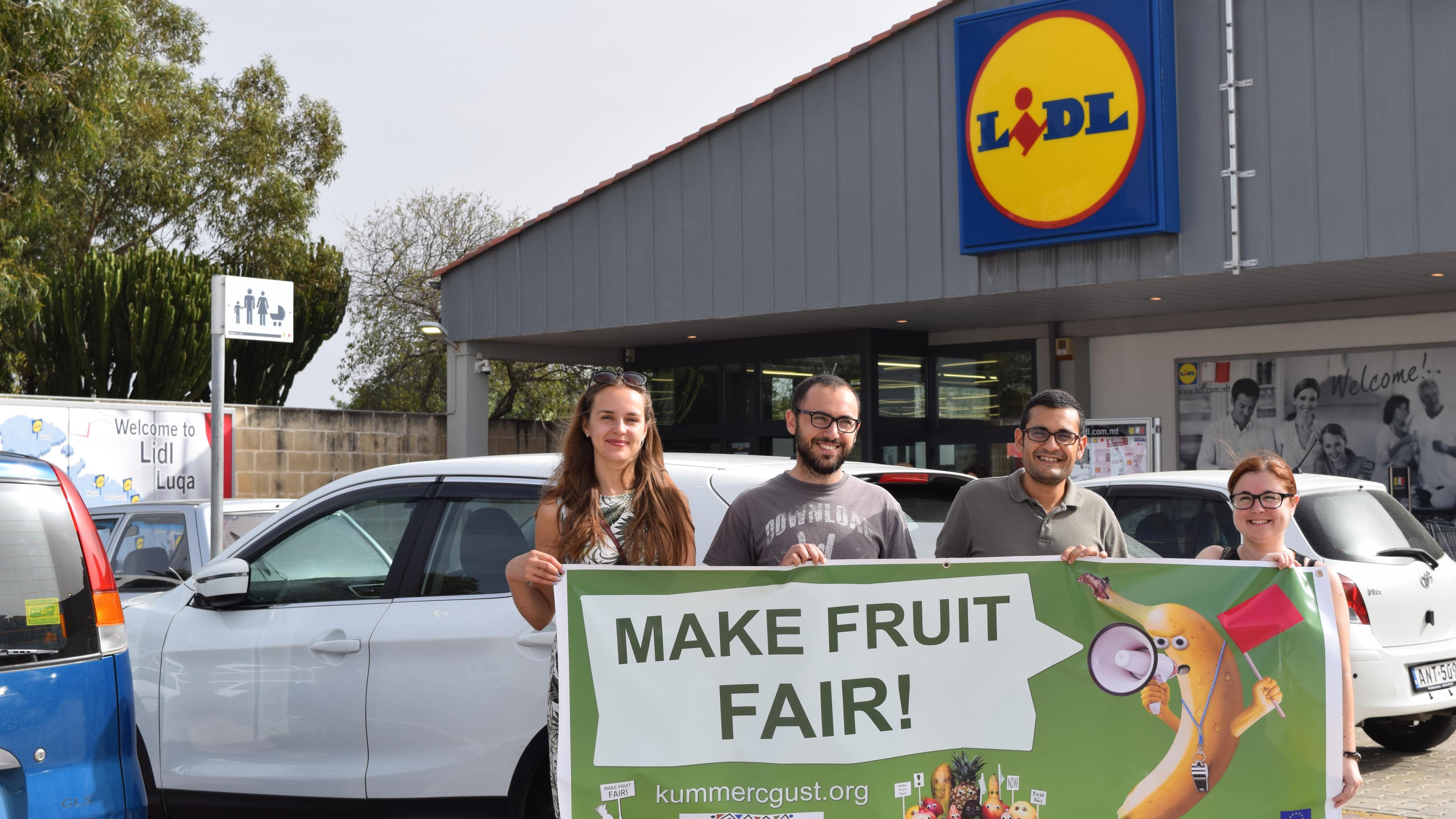 Aktivist/innen mit einem Transparent vor einer Lidl-Filiale. Auf dem Transparent fordert eine wütende Banane per Megafon: „Make Fruit Fair!“