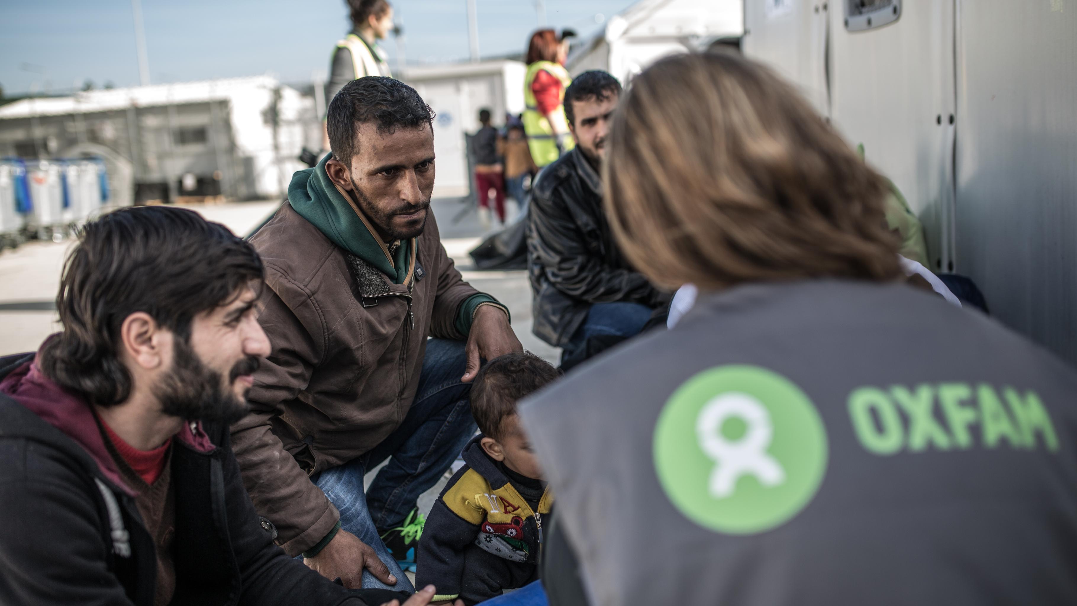 Eine Frau mit Oxfam-Weste, Rücken zur Kamera, spricht mit einer Familie in einem Flüchtlingscamp.