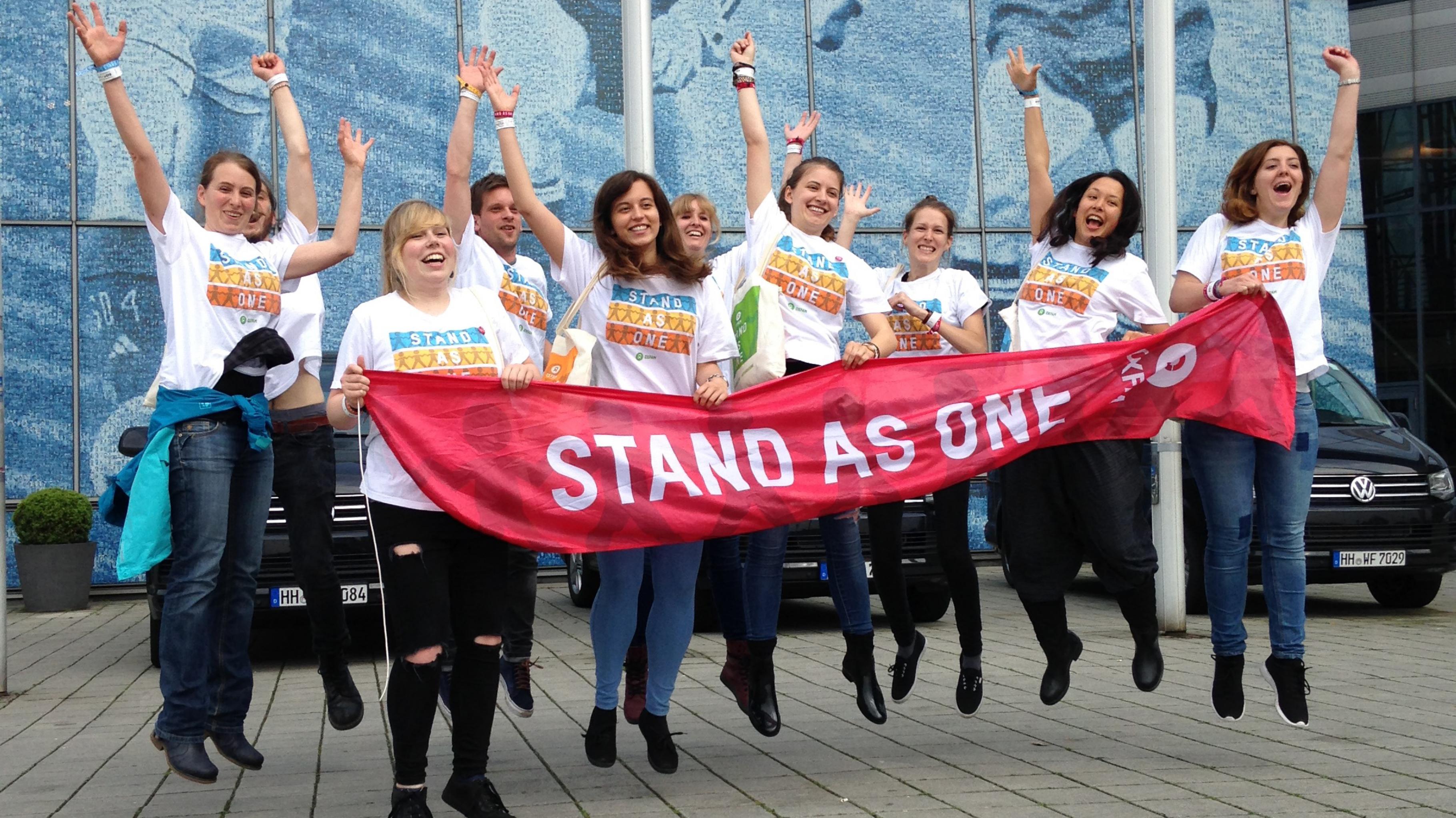 Hüpfende Konzertaktivistinnen halten ein Banner mit der Aufschrift: Stand As One