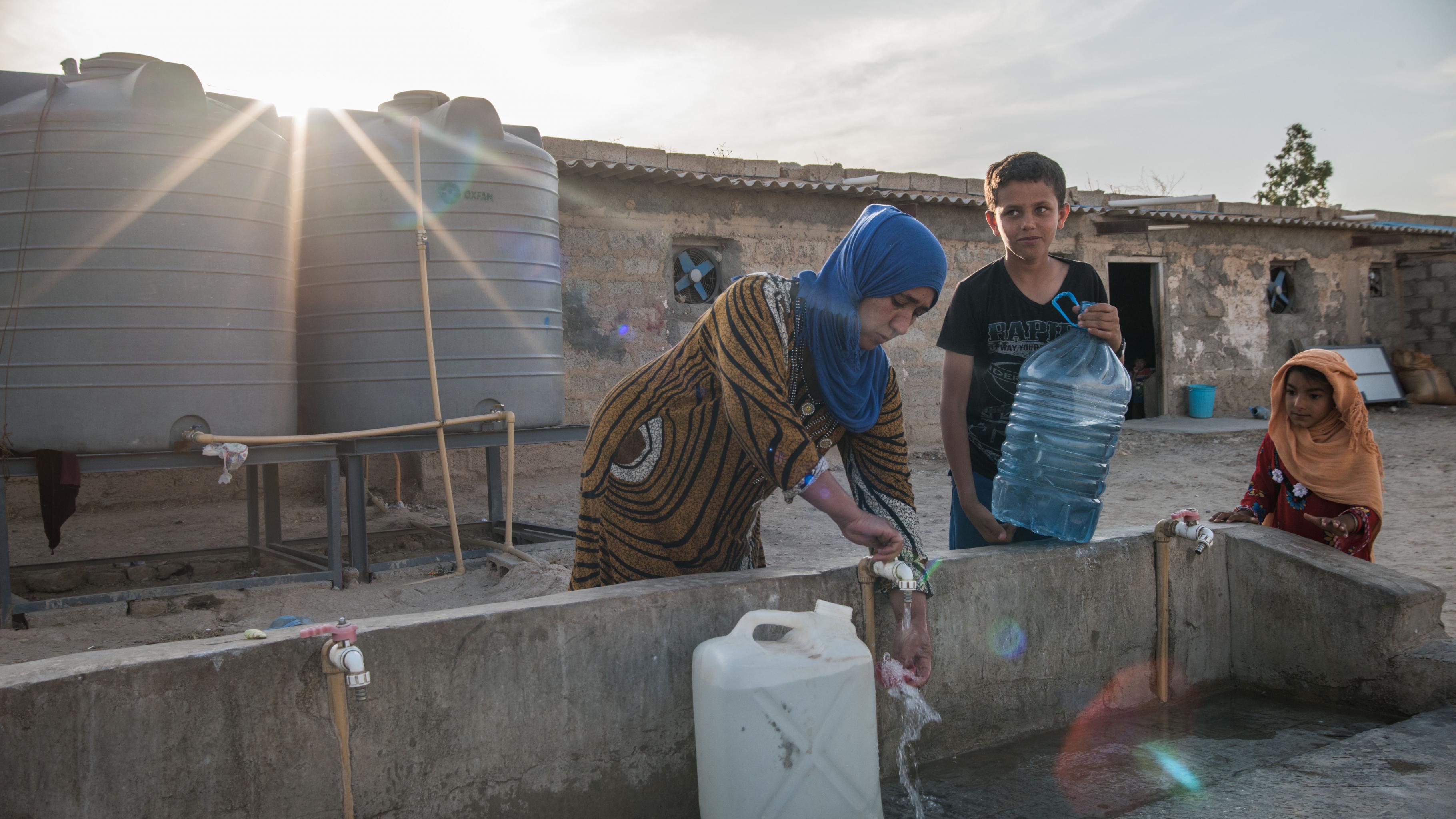 Eine Frau mit zwei Jungen füllt Wasserkanister an einem Wasserhahn auf. Im Hintergrund sind zwei Wassertanks und ein altes verlassenes Hauss zu sehen. 