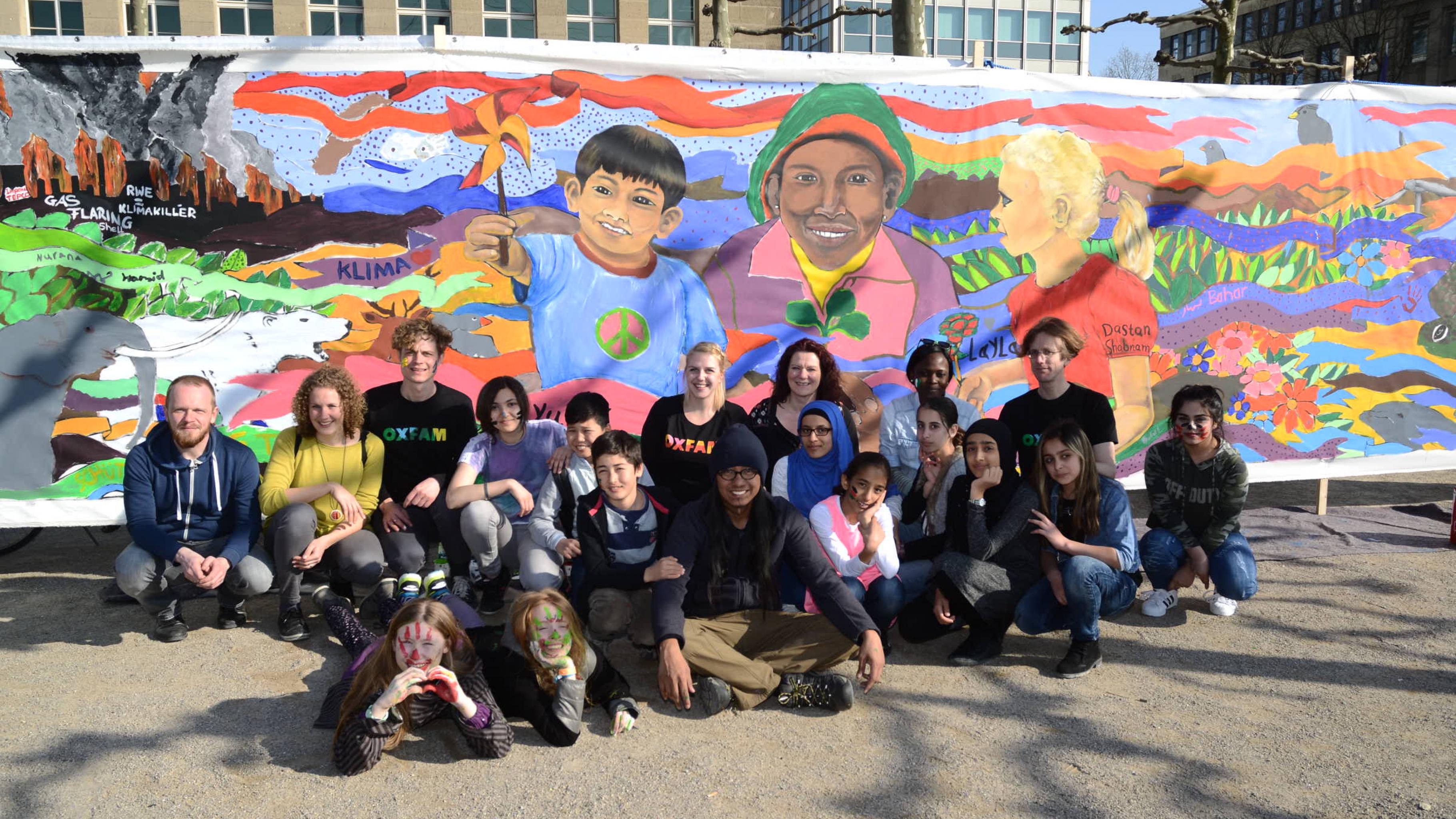 Künstler und Aktivist/innen vor dem fertigen Gemälde. Das Bild zeigt Kinder Asiens, Afrikas und Europas, die gemeinsam eine neue klimafreundliche Welt erschaffen.