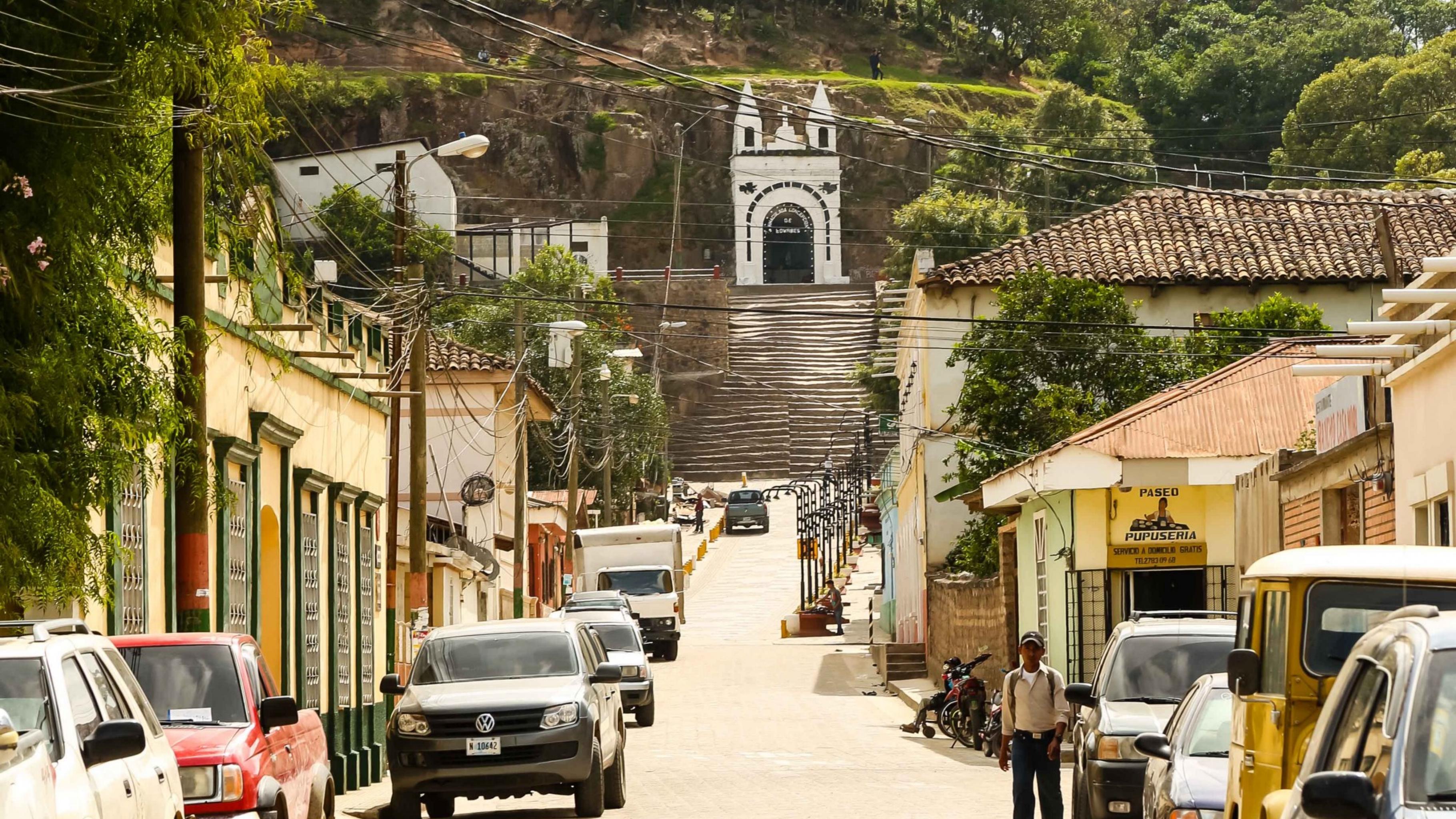 Eine sonnige kleine Straße, gesäumt von Autos und niedrigen Häusern, führt bergauf. Am Ende der Straße führt eine Treppe zu einer Kapelle an einem bewaldeten Berg.