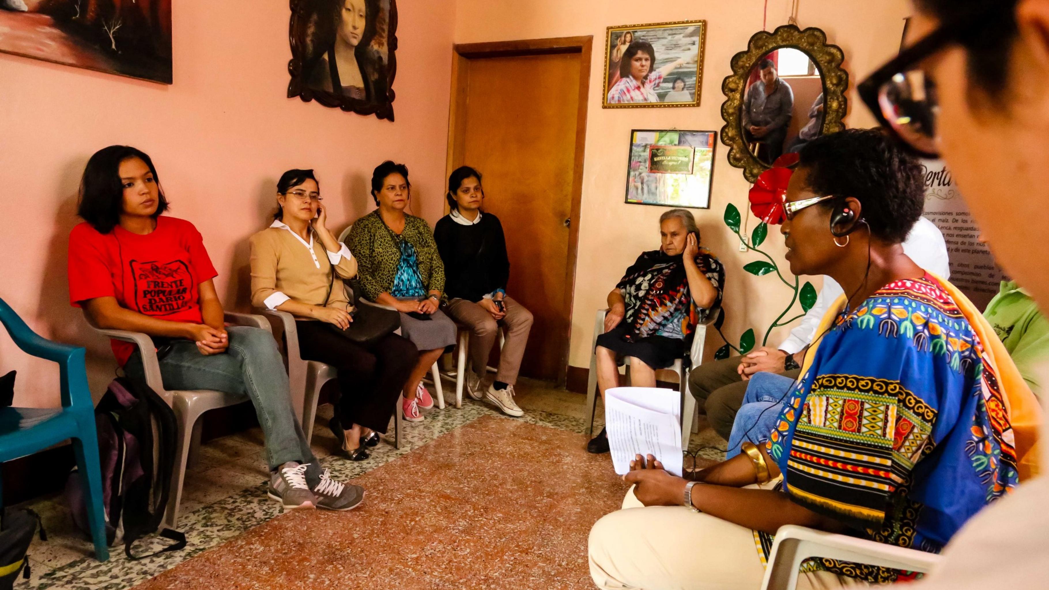In einem Zimmer sitzen Frauen und Männer auf Plastikstühlen im Kreis und sprechen miteinander. An den Wänden hängen Bilder, unter anderem von Berta Cáceres.