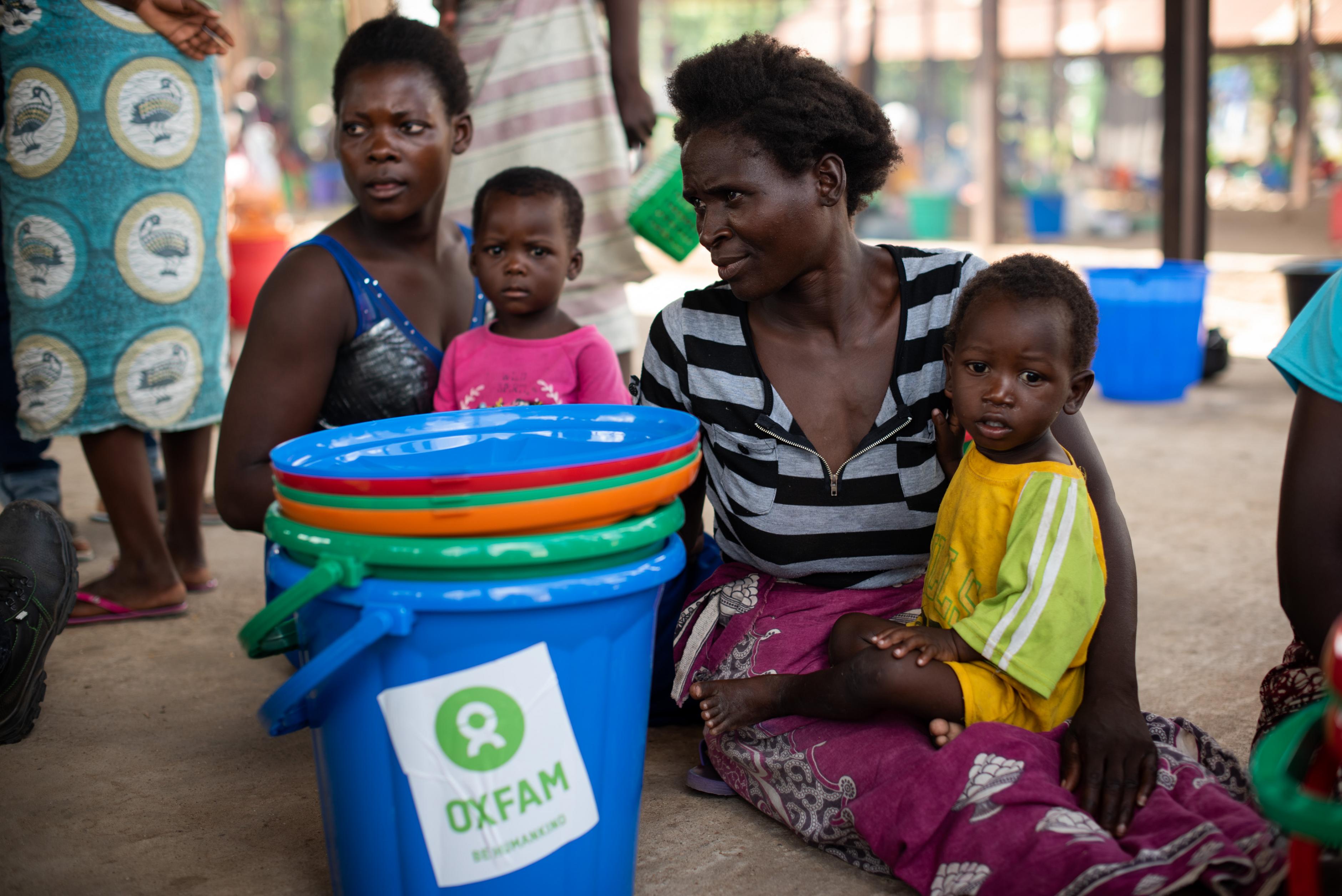 Zwei Frauen sitzen neben Oxfam-Eimern auf dem Boden, jeweils mit einem Kind im Schoß.