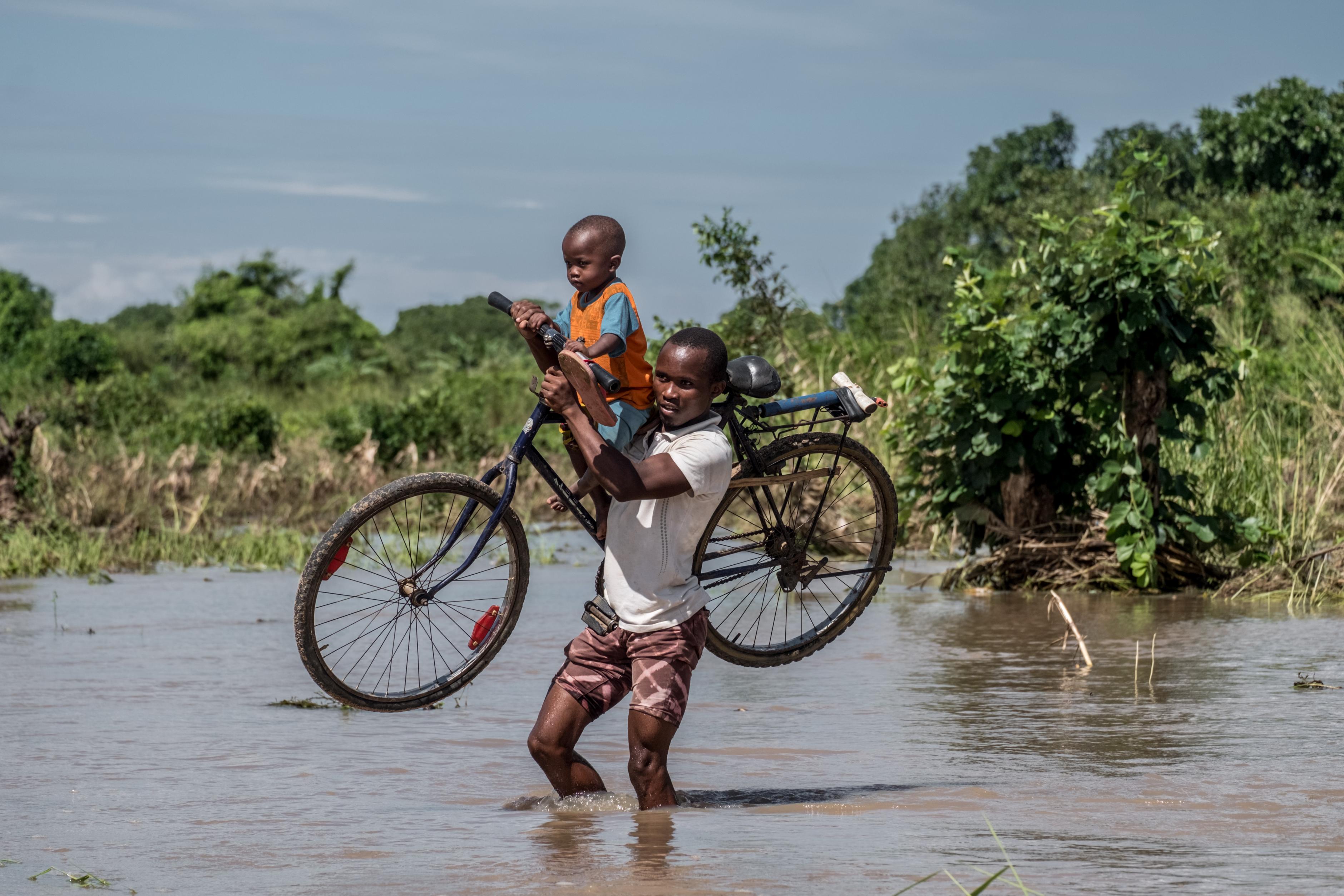 Ein Mann läuft durch Wassermassen während er ein Fahrrad und ein Kind trägt