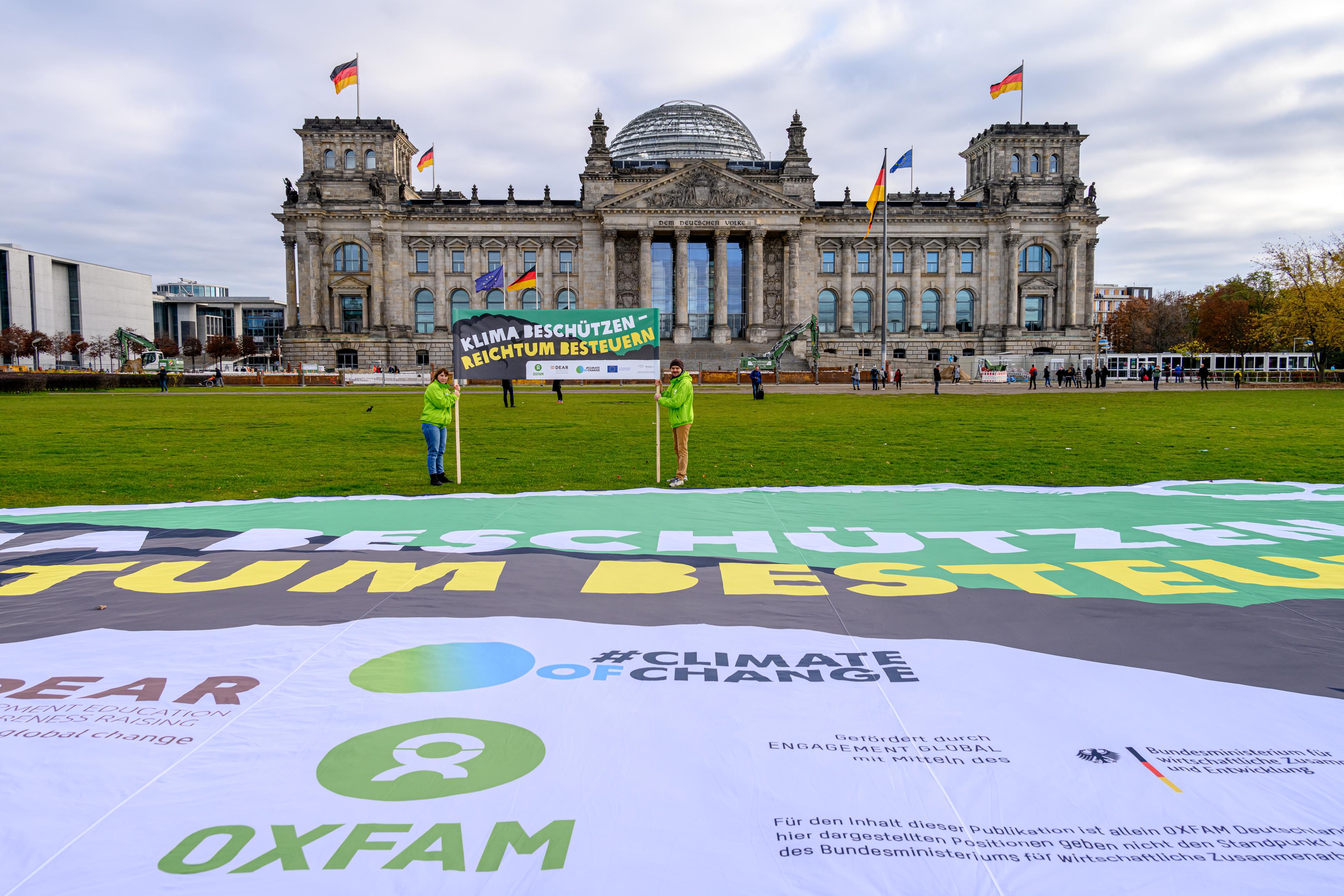 Oxfam-Aktivist*innen, mit grünen Jacken bekleidet, stehen vor dem Reichstag und halten ein Banner mit der Aufschrift "Klima schützen - Reichtum besteuern" zwischen ihnen. Vor ihnen auf der Wiese befindet sich ein überdemensionales Transparent auf der Wiese.
