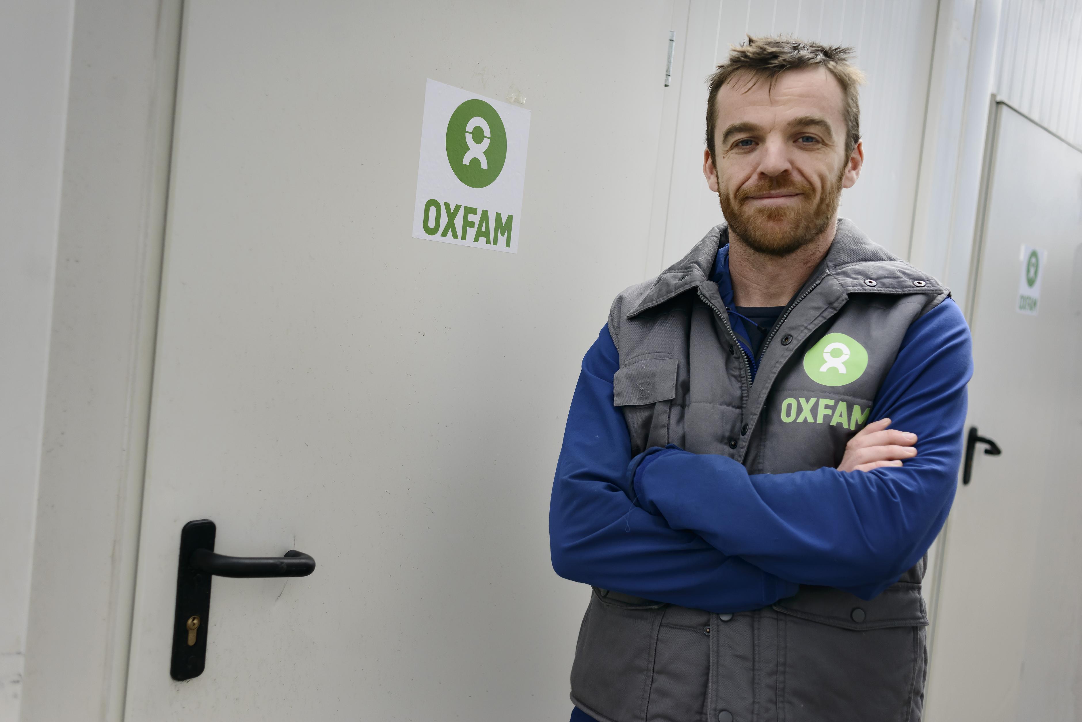 Angus Mc Bride steht vor einer Dusche mit dem Oxfam-Symbol auf der Tür. Er trägt einen blauen Pullover und eine graue Oxfam-Weste darüber.