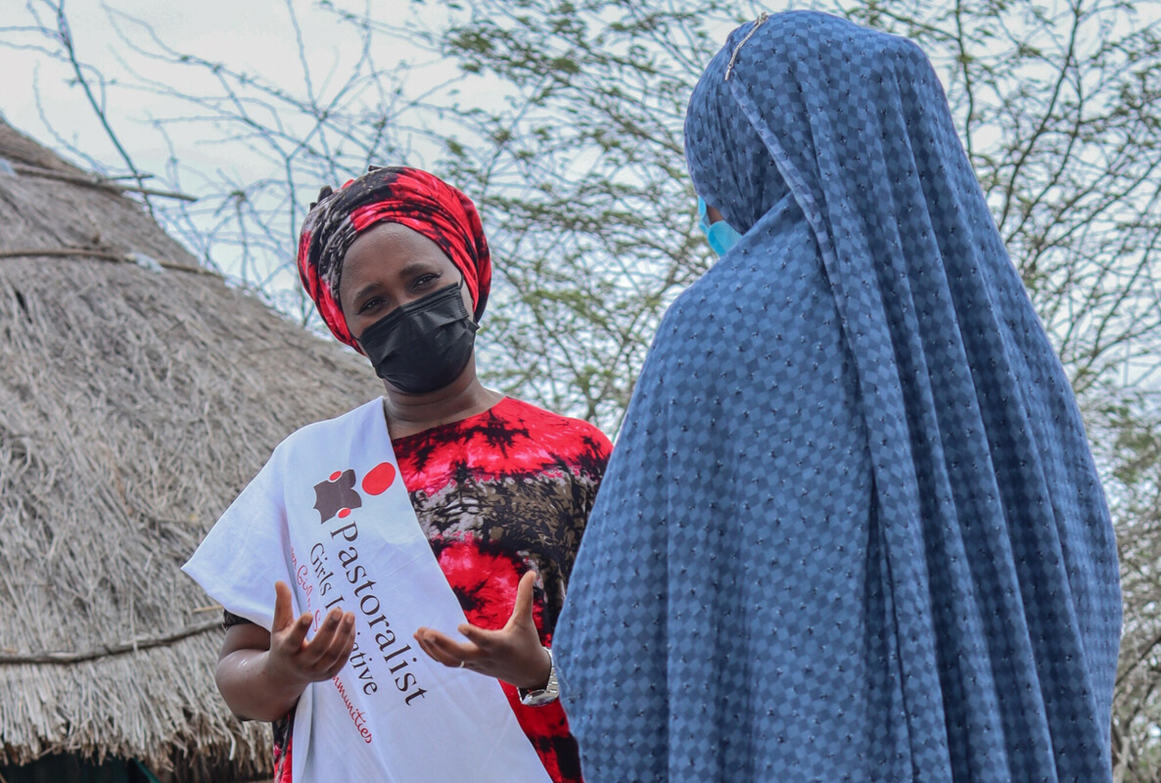 Margaret Kikuvi trägt eine medizinische Maske und eine Schärpe mit der Aufschrift „Pastoralist Girls Initiative“. Sie spricht mit einer zweiten Frau.