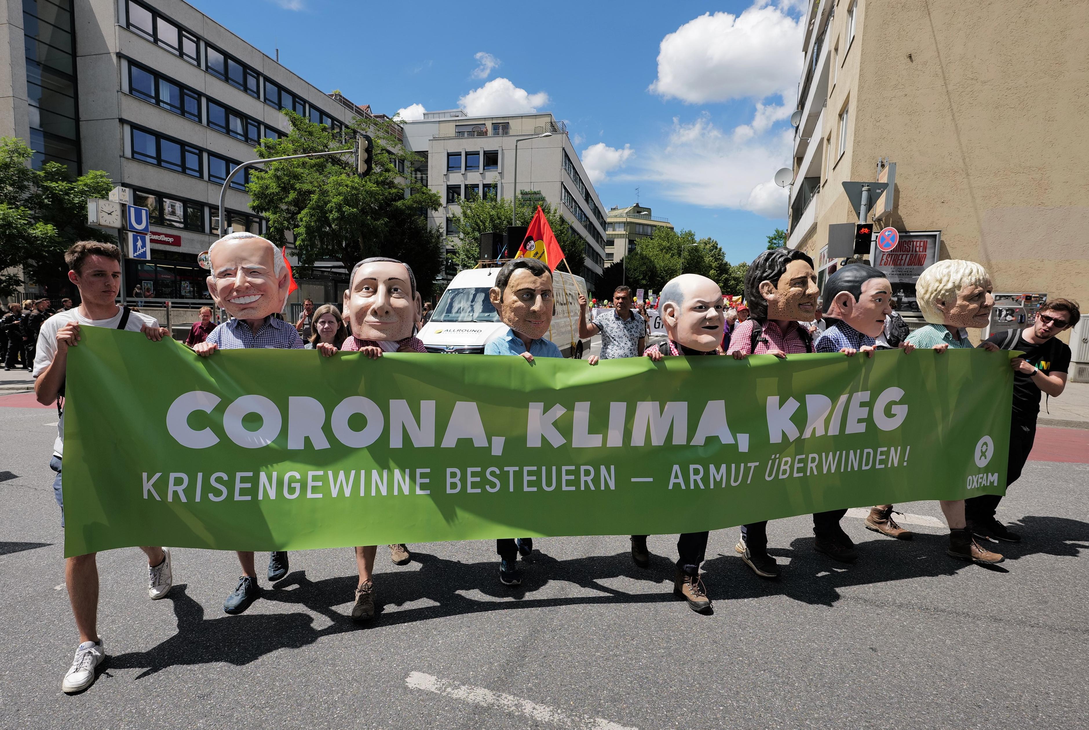 9 Oxfam-Mitarbeiter*innen tragen einen grünen Banner mit der Aufschrift "Corona, Klima, Krieg: Krisengewinne besteuern - Armut überwinden" auf der G7-Demo 2022. 7 von ihnen (mittig) tragen Big-Heads und posieren so als G7-Regierungschefs.