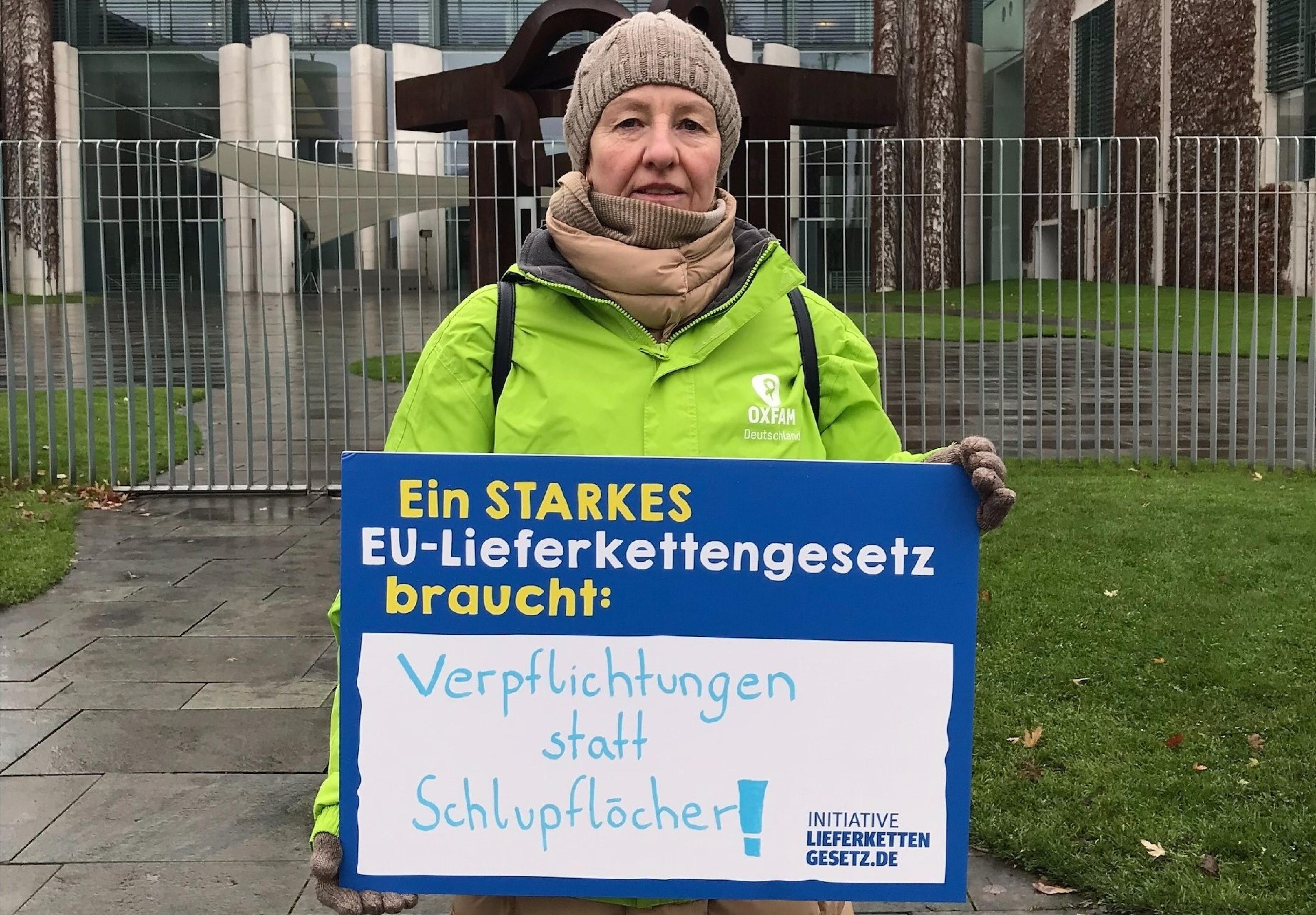 Oxfam-Ehrenamtliche, in grüner Oxfam-Jacke, vor dem Kanzleramt mit einem Schild der Initiative Lieferkettengesetz. Aufschrift: "Ein starkes EU-Lieferkettengesetz braucht: Verpflichtungen statt Schlupflöcher"