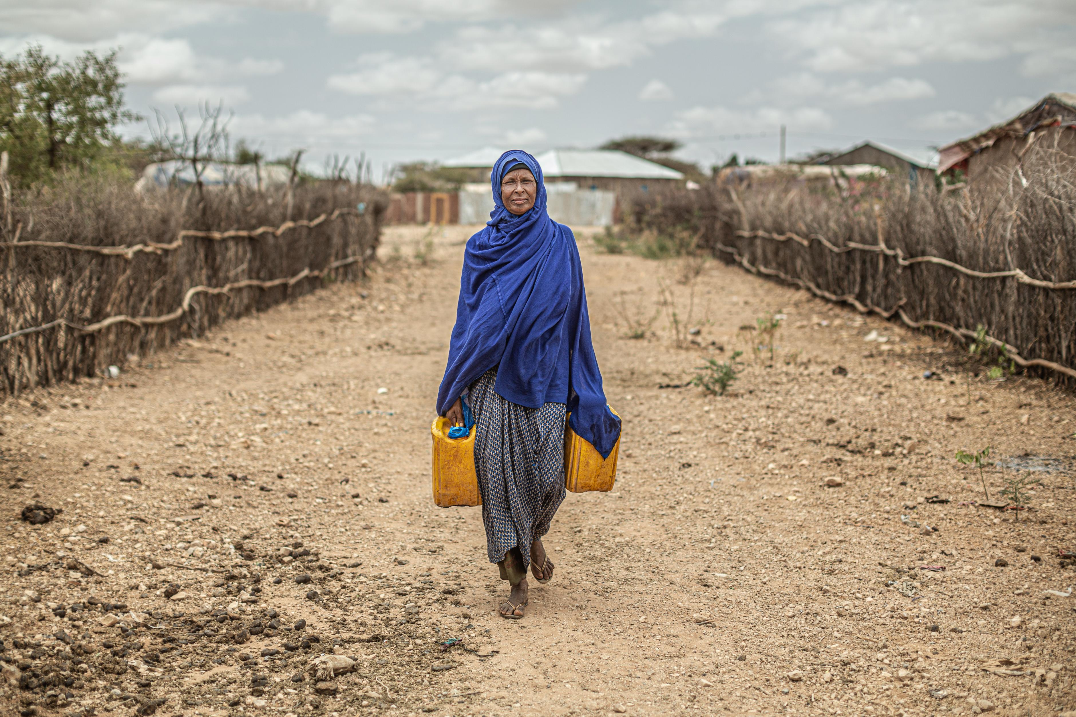 Amina Ibrahim läuft einen Sandweg entlang, der von Zäunen - bestehend aus Weidenästen - begrenzt ist. In beiden Händen hält sie gelbe Wasser-Kanister.