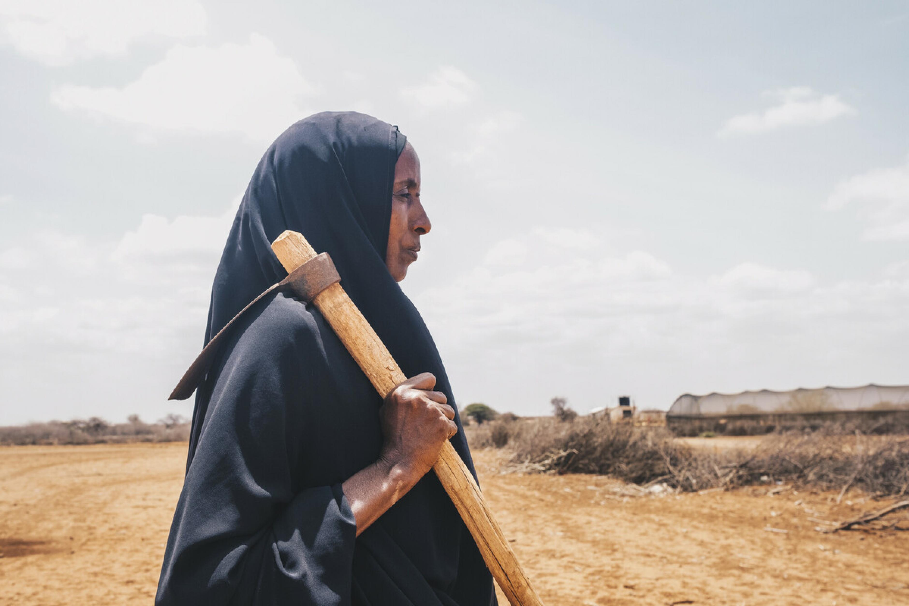 Diyaara* steht auf dem Acker in Wajir, Kenia, der früher ihre Familie ernährt hat. Sie trägt eine Hacke. In Ostafrika herrscht eine massive Dürre, aus der eine Hungerkrise entstanden ist. Die steigenden Getreidepreise verschlimmern die Lage zusätzlich.