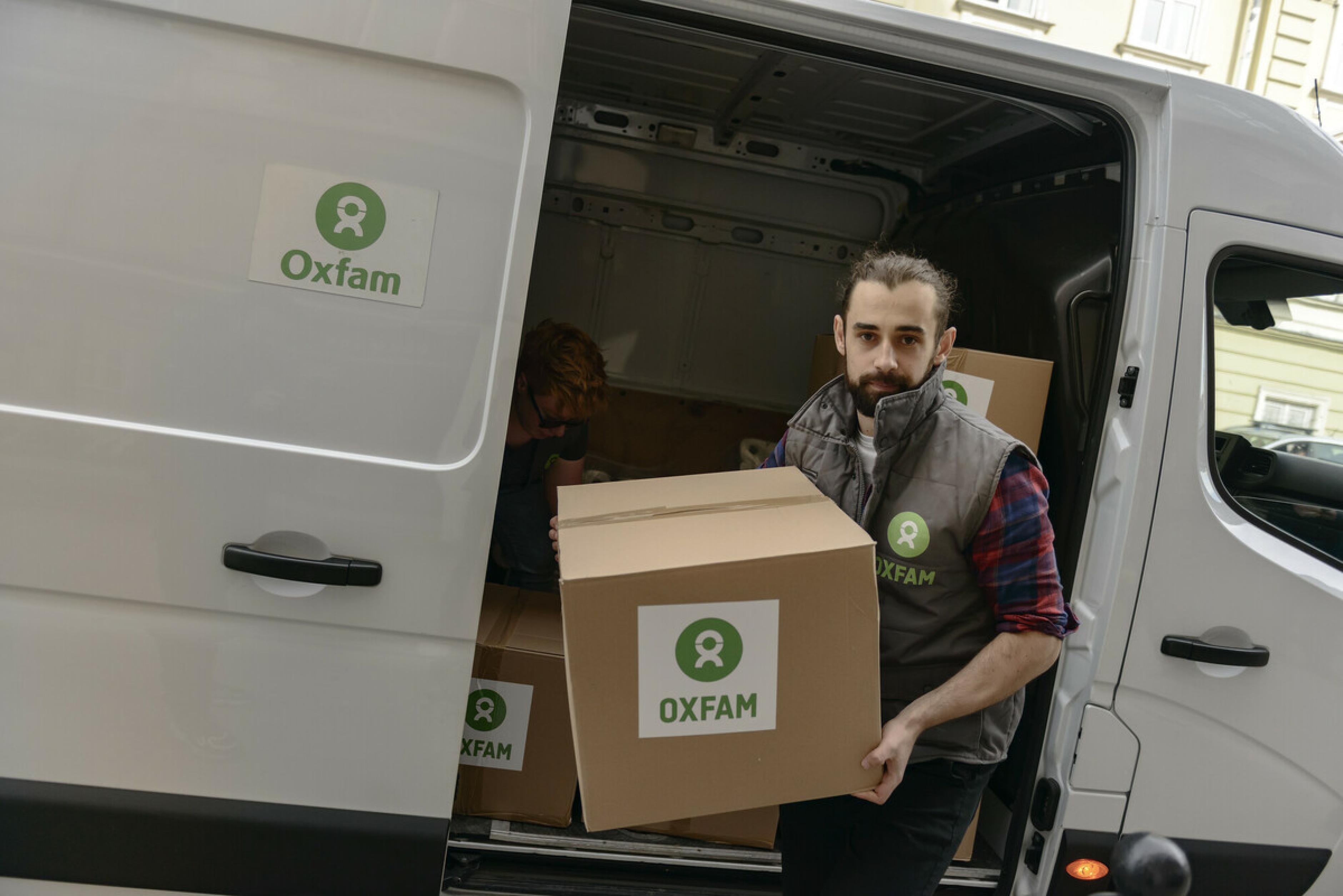 Ein Mann in Oxfam-Weste trägt einen Karton aus einem weißen Transporter. Auf dem Karton ist ein Oxfam-Aufkleber.